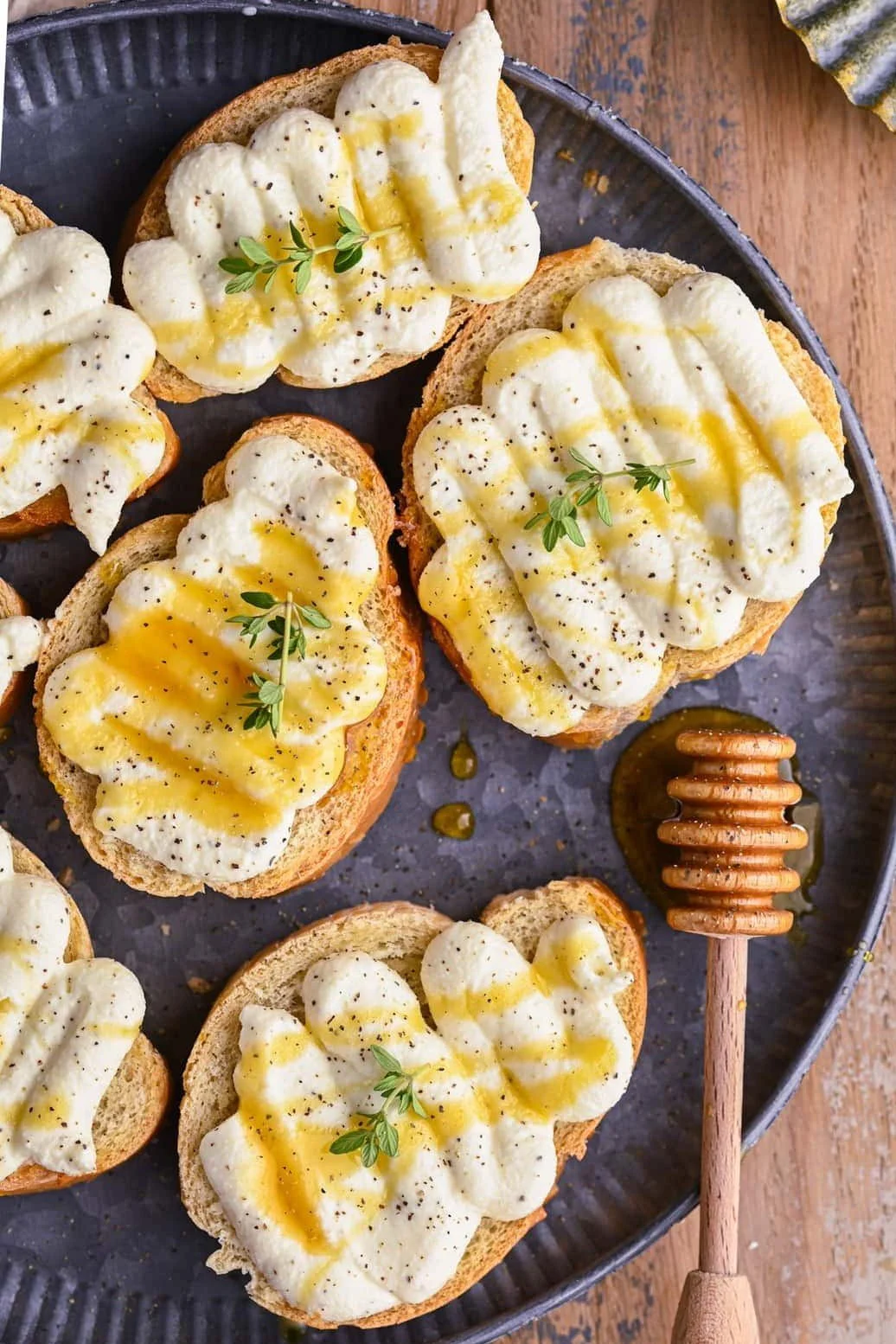 Slice of toasted bread topped with creamy mashed potatoes, drizzled with honey, garnished with fresh herbs, and sprinkled with black pepper on a dark round tray, with a honey dipper beside it.