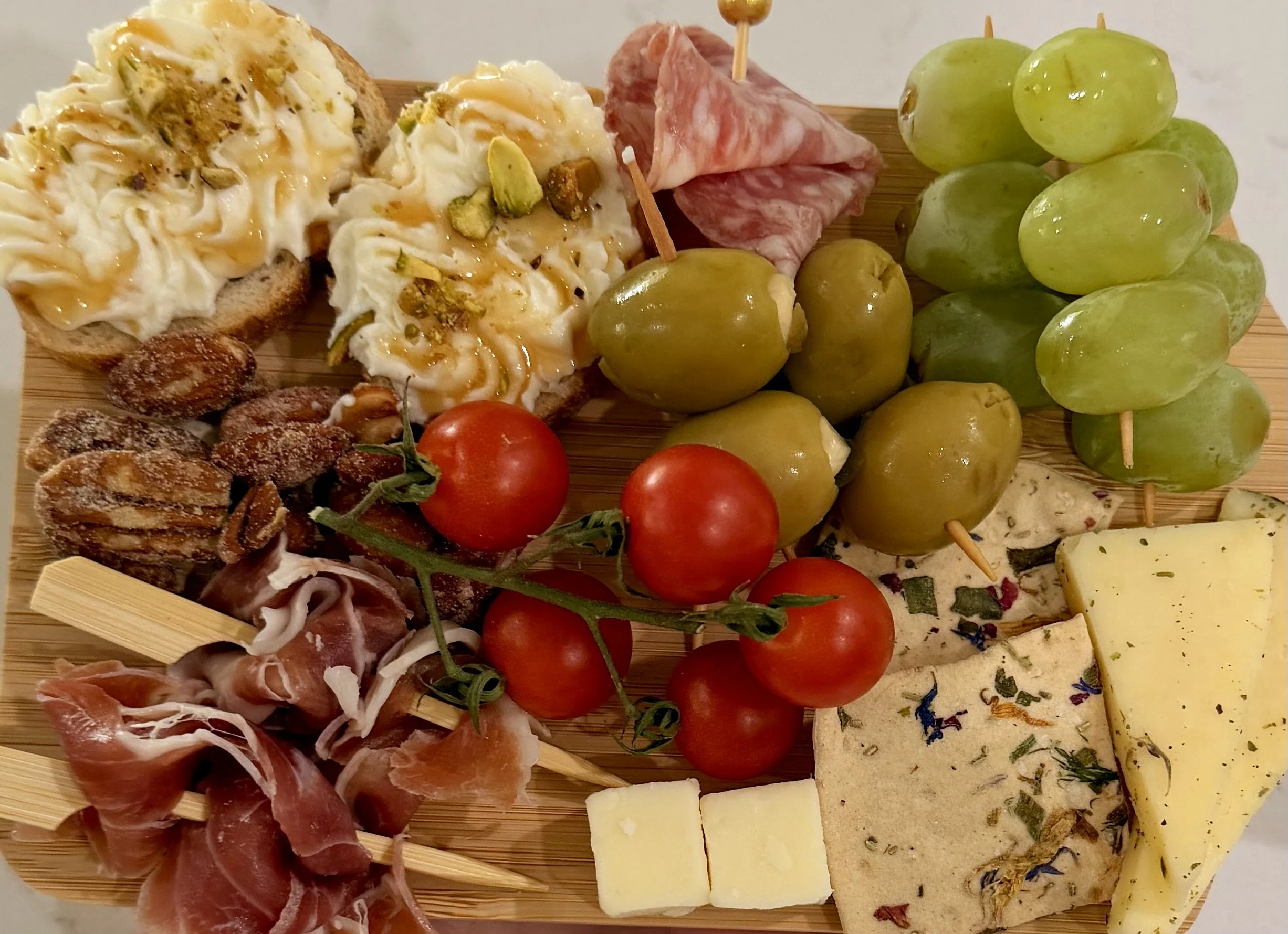 A cheese and charcuterie platter with green grapes, cherry tomatoes, prosciutto, salami, cheese slices, a slice of herb cheese, and bread topped with cheese and nuts.