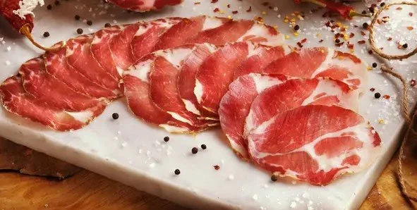 Thinly sliced cured meat, likely prosciutto, arranged on a white platter with some black peppercorns and festive gold and white decorations.