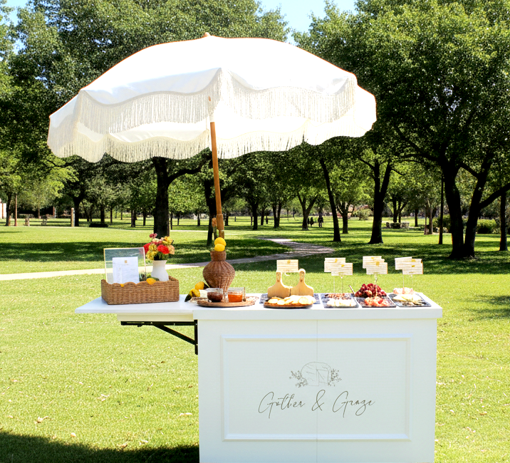 A white outdoor dessert table with a large white umbrella in a park, featuring a basket of flowers, a pitcher, and various desserts like cheese and fruit, with trees in the background.