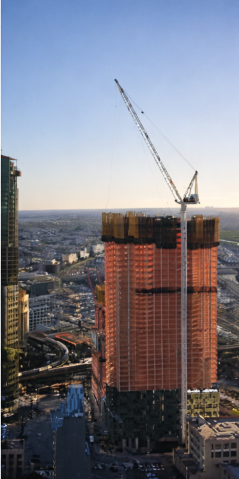 A high-rise building under construction with a tower crane on top, set against a cityscape background during sunset.