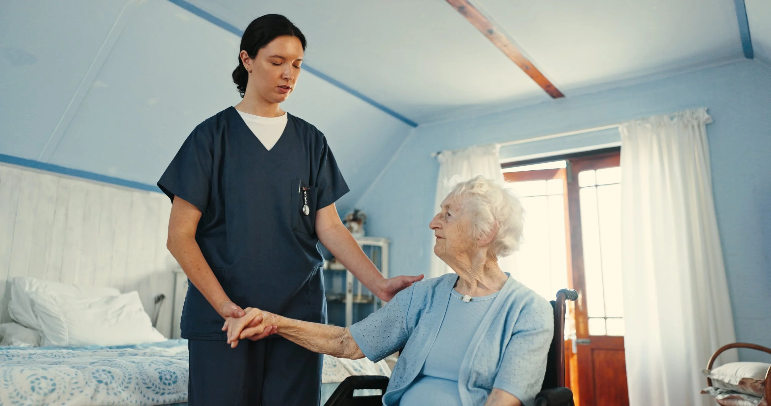Caregiver holding hands with a senior woman in a wheelchair in her room.