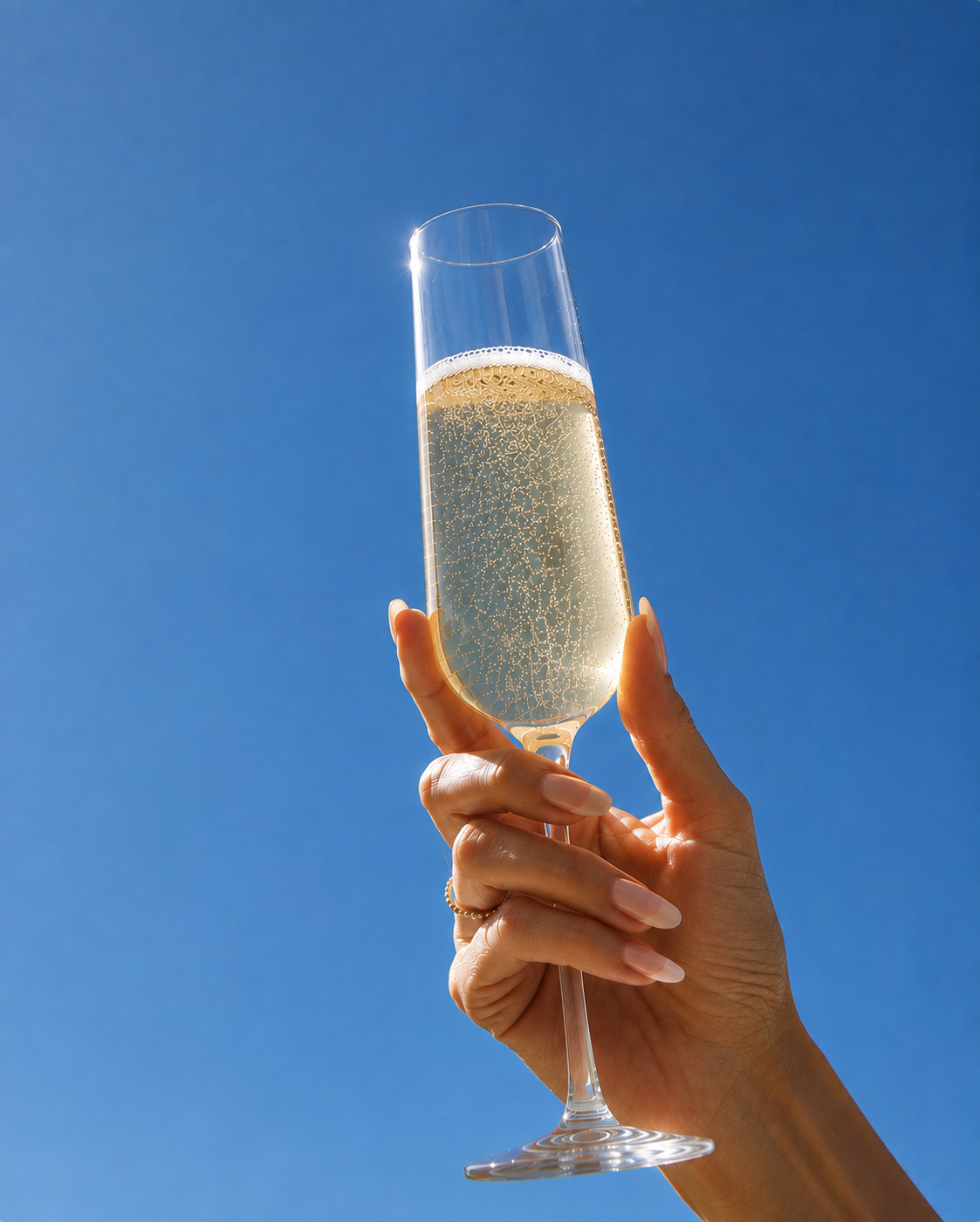 A hand holding a glass of IYLIA Premium Gold Sparkling Non-Alcoholic wine against a clear blue sky.