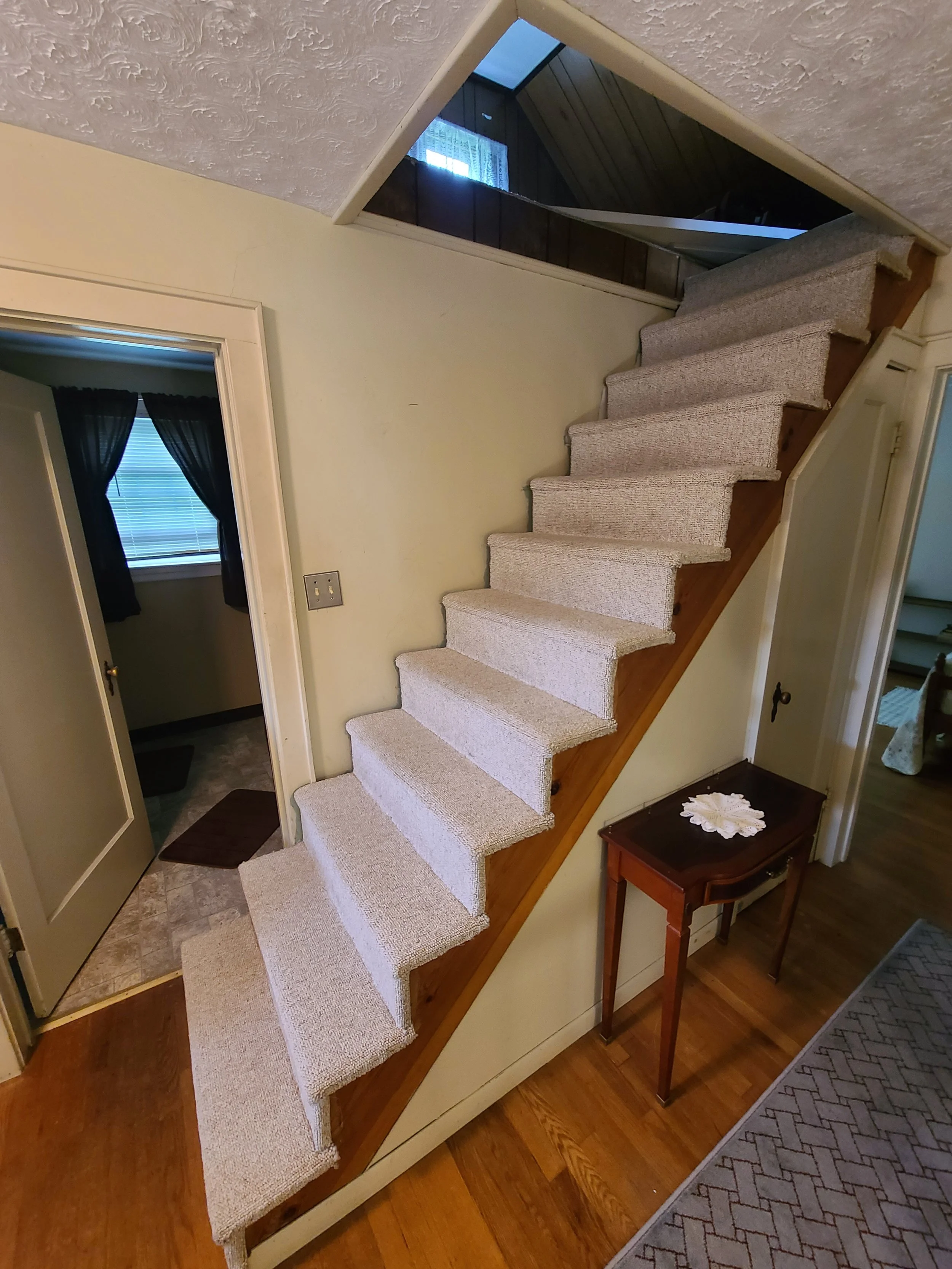 Interior view of a staircase with beige carpeting, leading from a small landing to the upper floor. The staircase is adjacent to a pale yellow wall and a small wooden table with a doily. A mirror above the stairs reflects the ceiling and window of the upper floor.