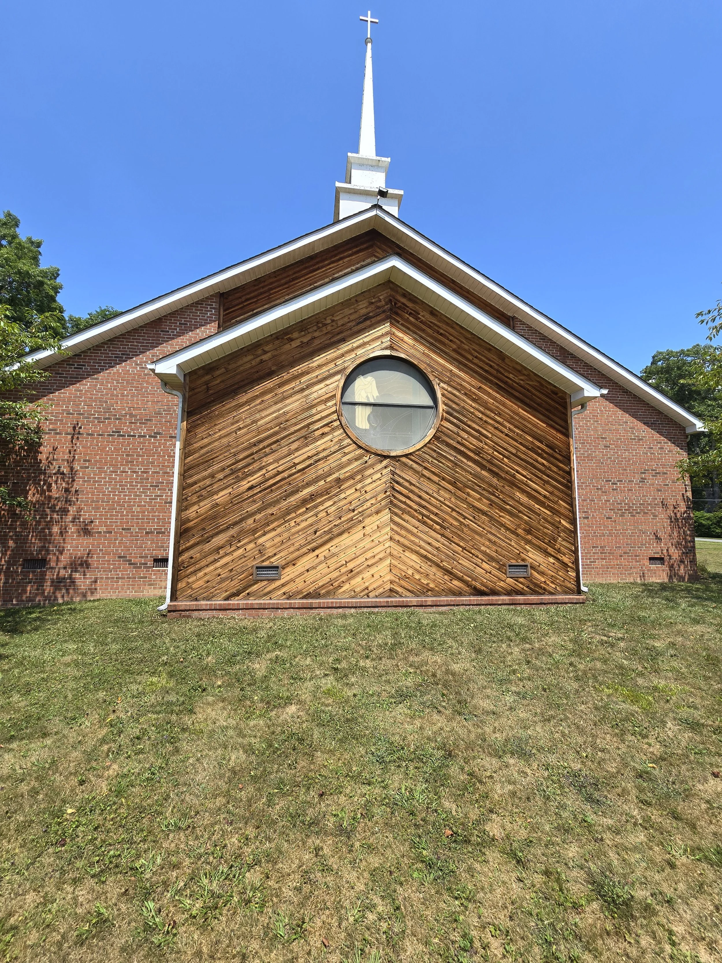 Front view of a church with a brick exterior and wooden front facade, circular stained glass window, and a white steeple with a cross on top, set against a bright blue sky.