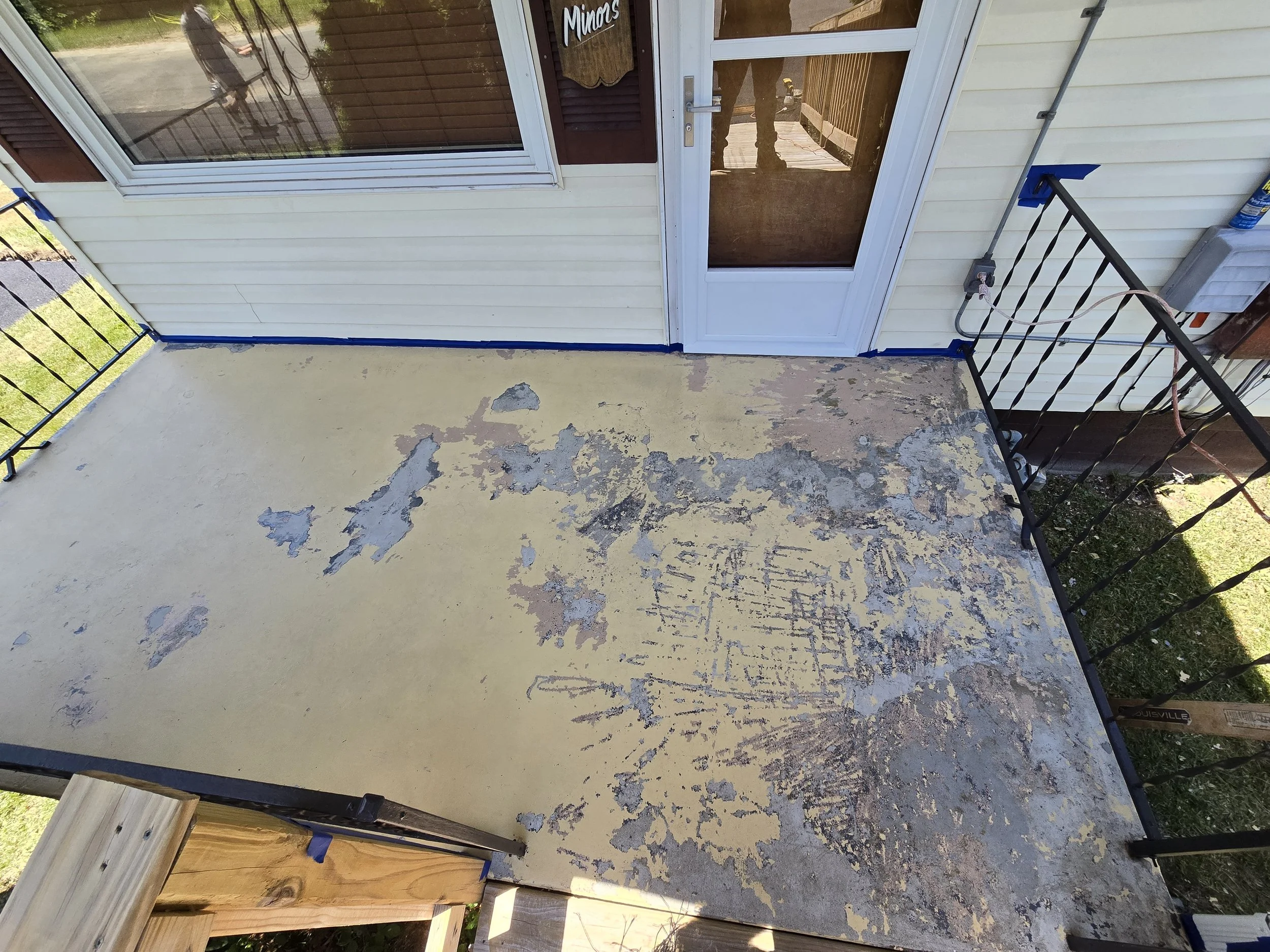 A weathered porch with peeling yellow paint revealing gray and brown underneath, surrounded by a black metal railing, with a door and a window on the house wall.