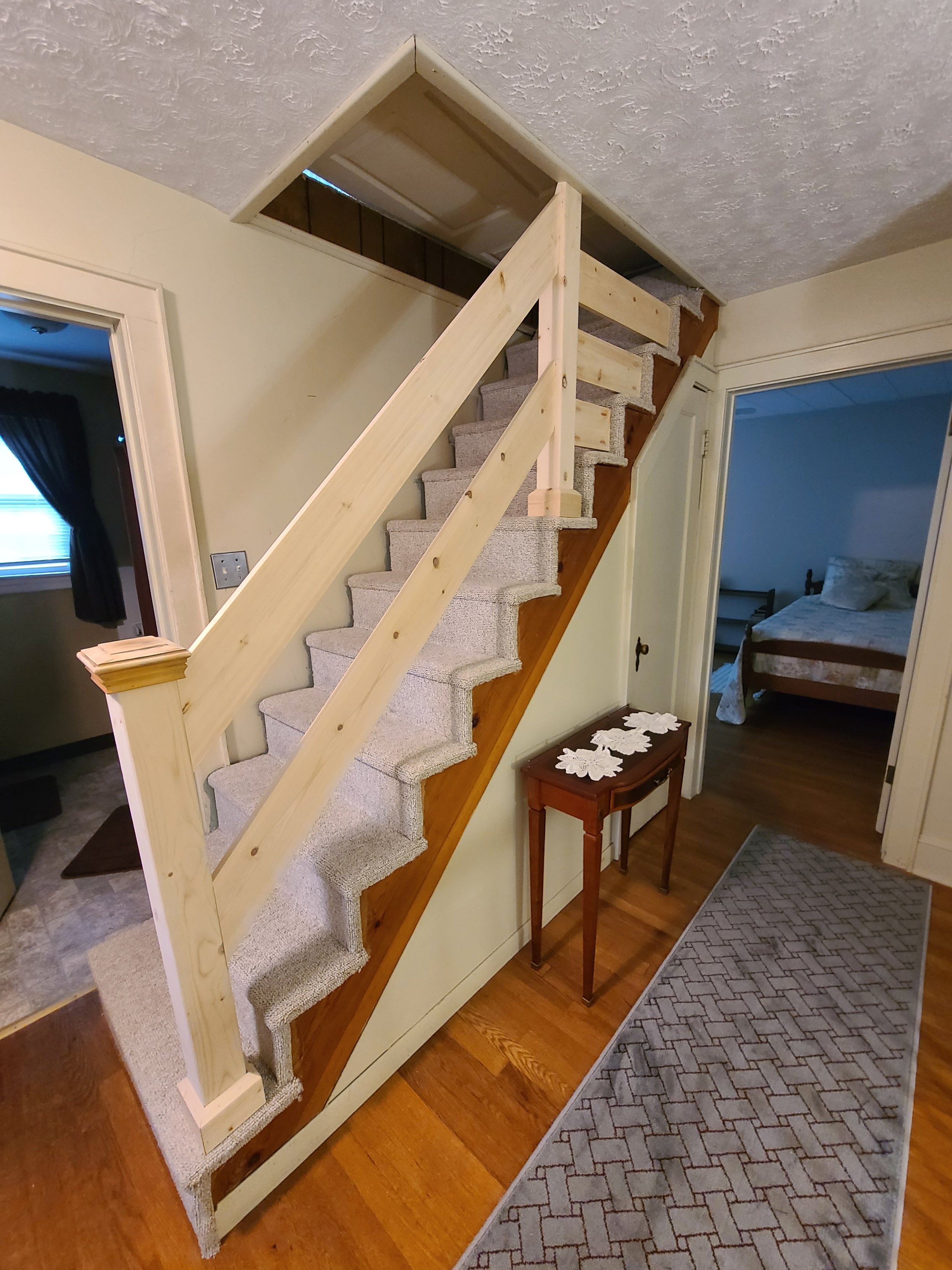 Interior view of a staircase with unfinished light wood railing, leading to an attic or second floor, with a small wooden table and a patterned runner rug on a hardwood floor, and a doorway to a bedroom.