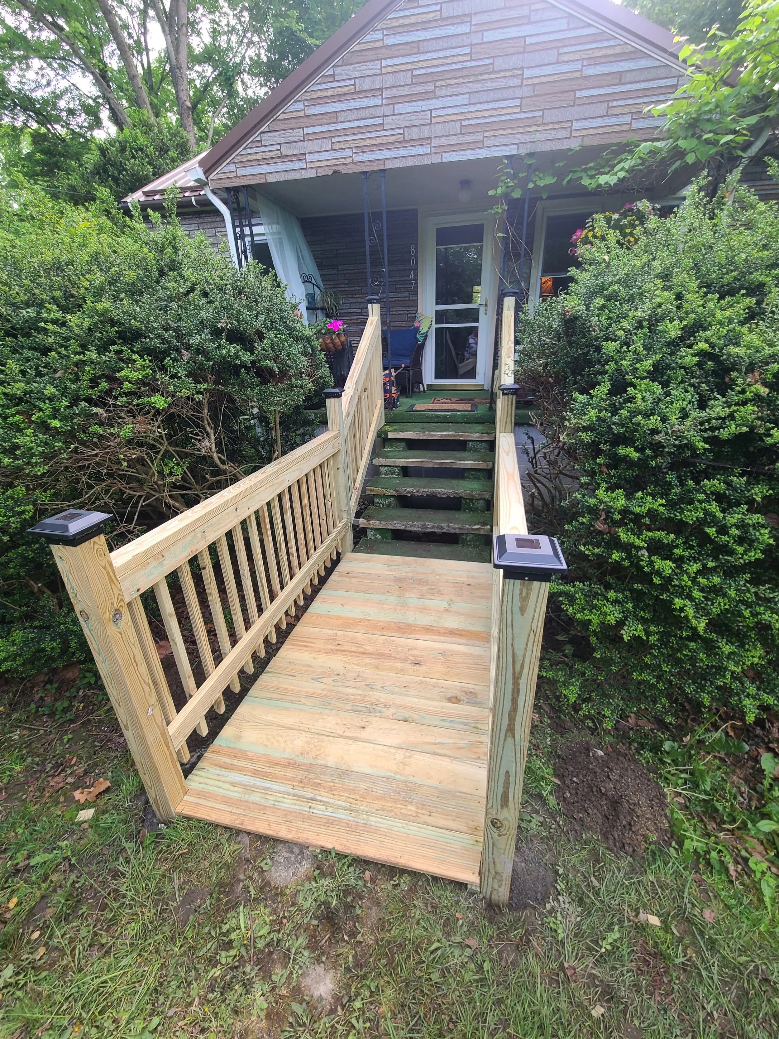 Front view of a house with a newly built wooden ramp and stairs leading up to the front door, surrounded by greenery and bushes.