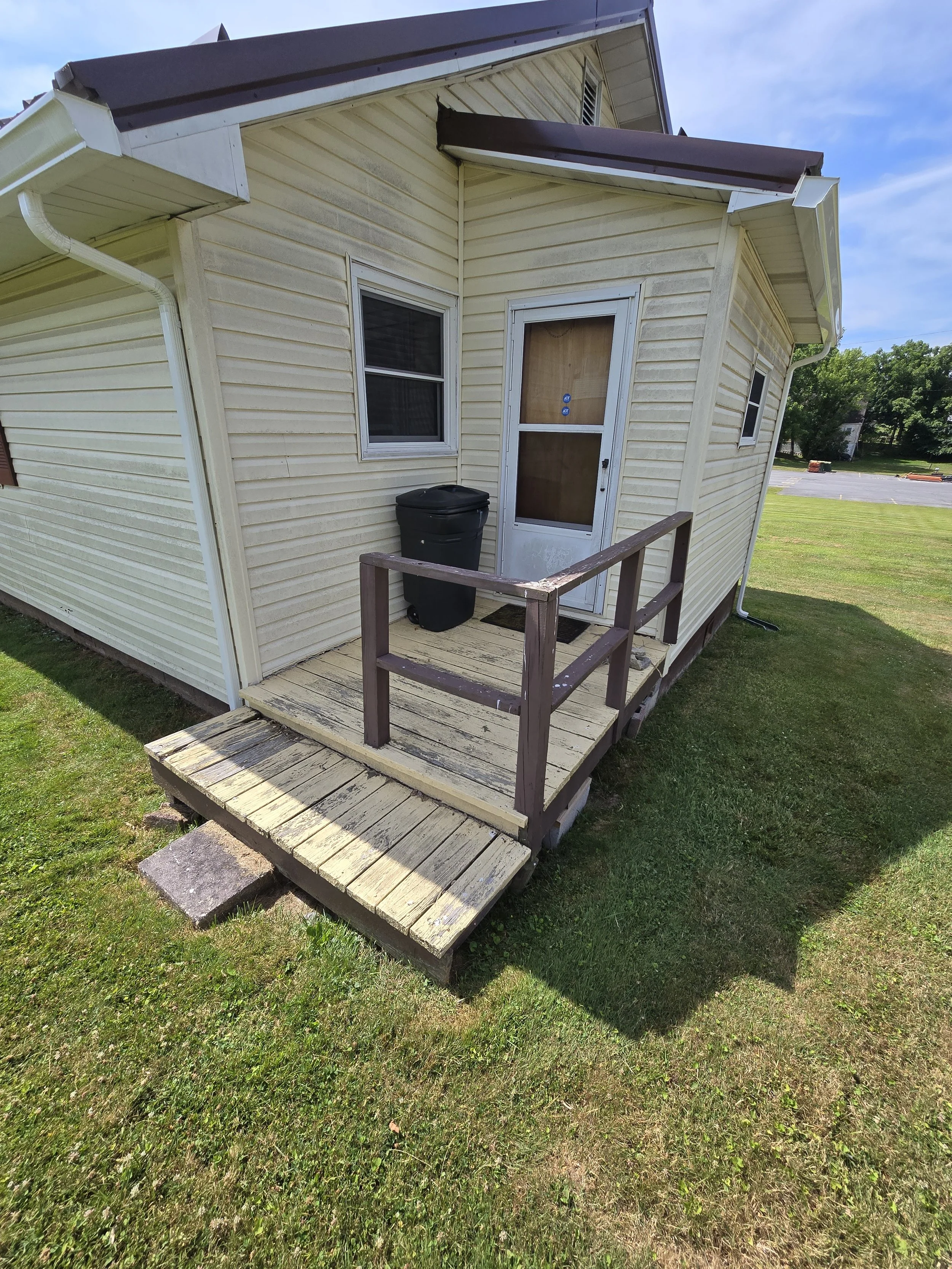 A small beige house with a wooden ramp and railing in front of the door, a black trash can, two small windows, and a sloped brown roof, with a grassy yard and parking lot in the background.