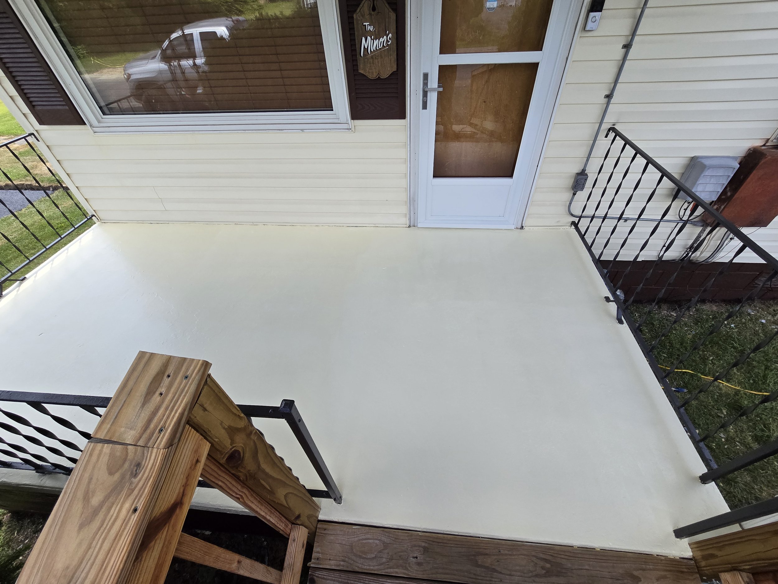 Newly painted white concrete porch with black metal railing, beige siding, and a glass door leading inside. A wooden staircase with railing starts at the bottom of the image. A window with blinds reflects a parked car outside.