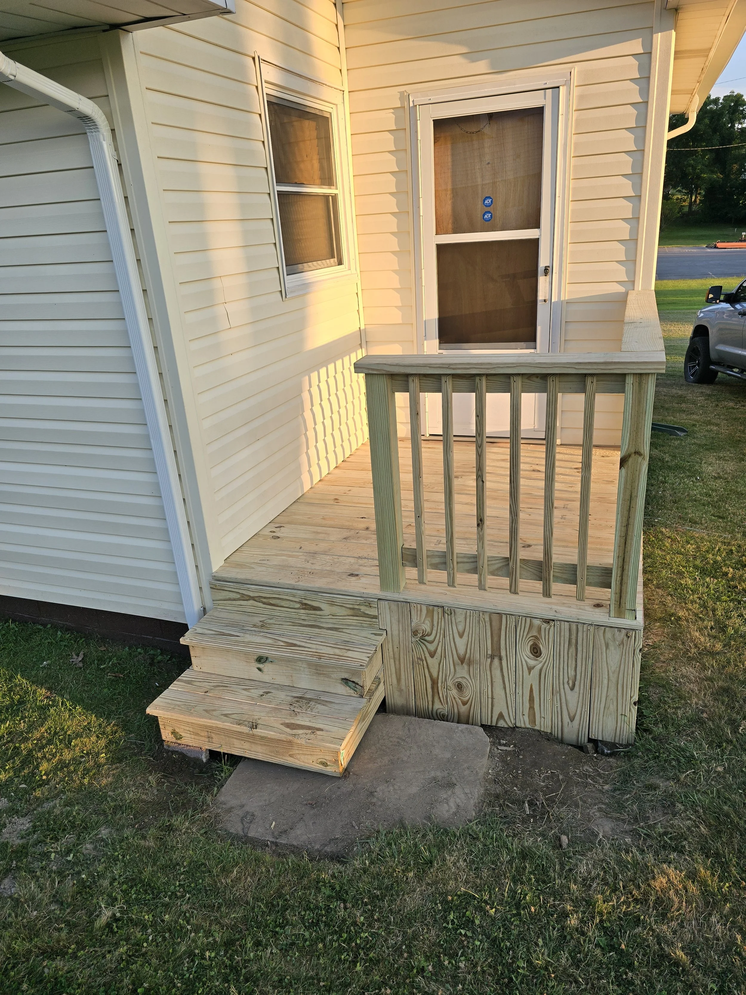 Newly built small wooden porch with a railing and steps attached to the side of a house, with a door and window visible.