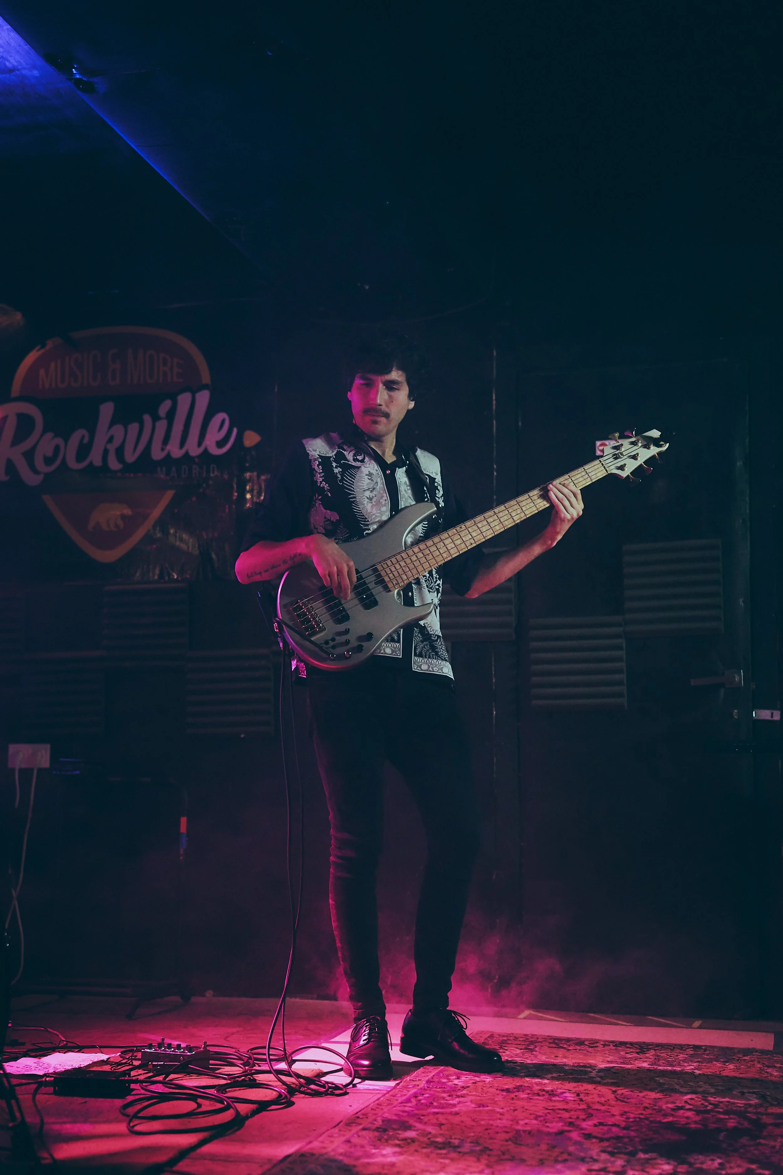 A musician playing an electric bass guitar on stage at Rockville Madrid, under pink and purple lighting, with cables and pedals on the floor, and a dark background with a logo that says 'Music & More Rockville Madrid'.