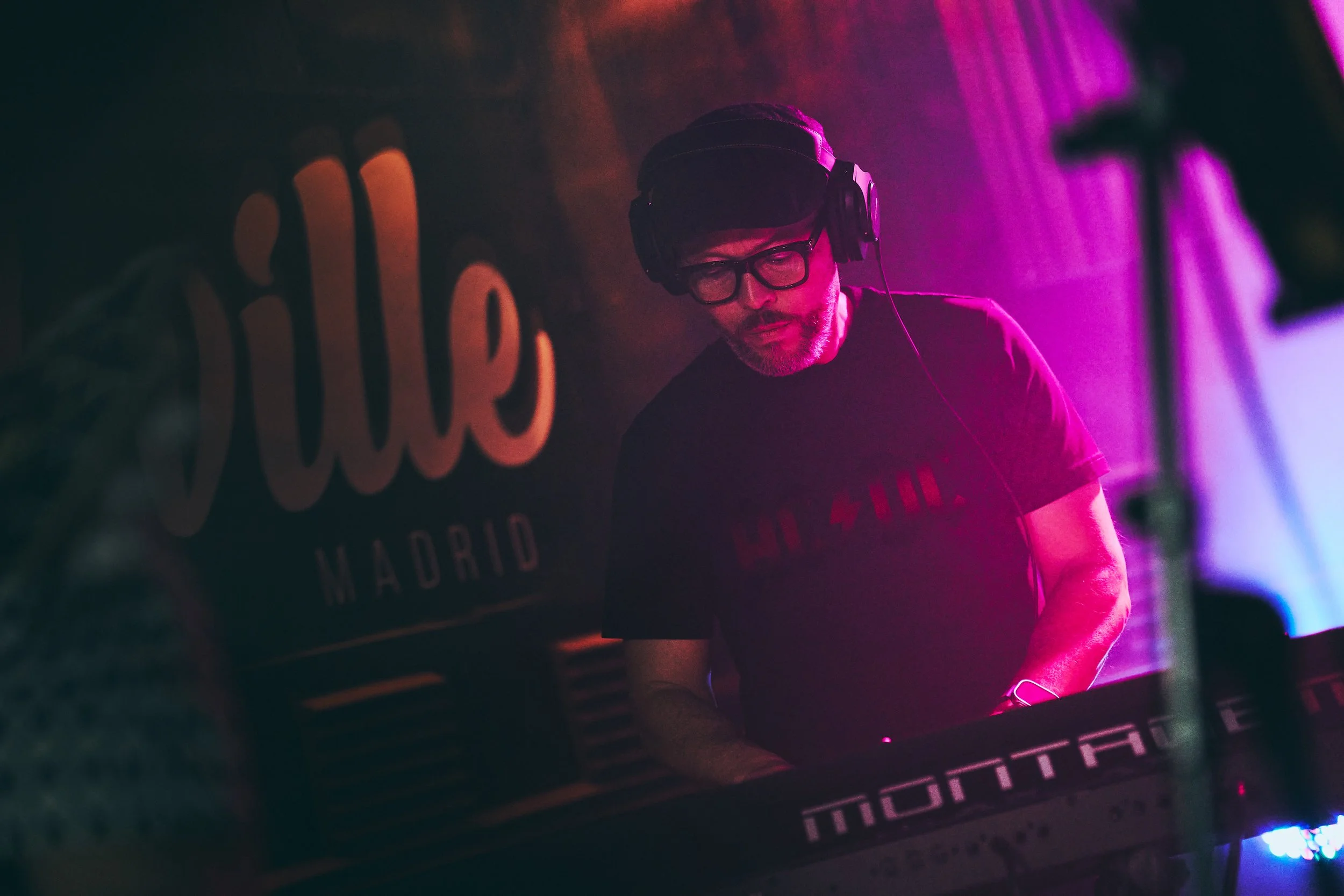 A DJ wearing glasses and headphones is playing music at a nightclub or event with colorful pink, purple, and blue lighting. There is a large sign that says "Cube Madrid" in the background.