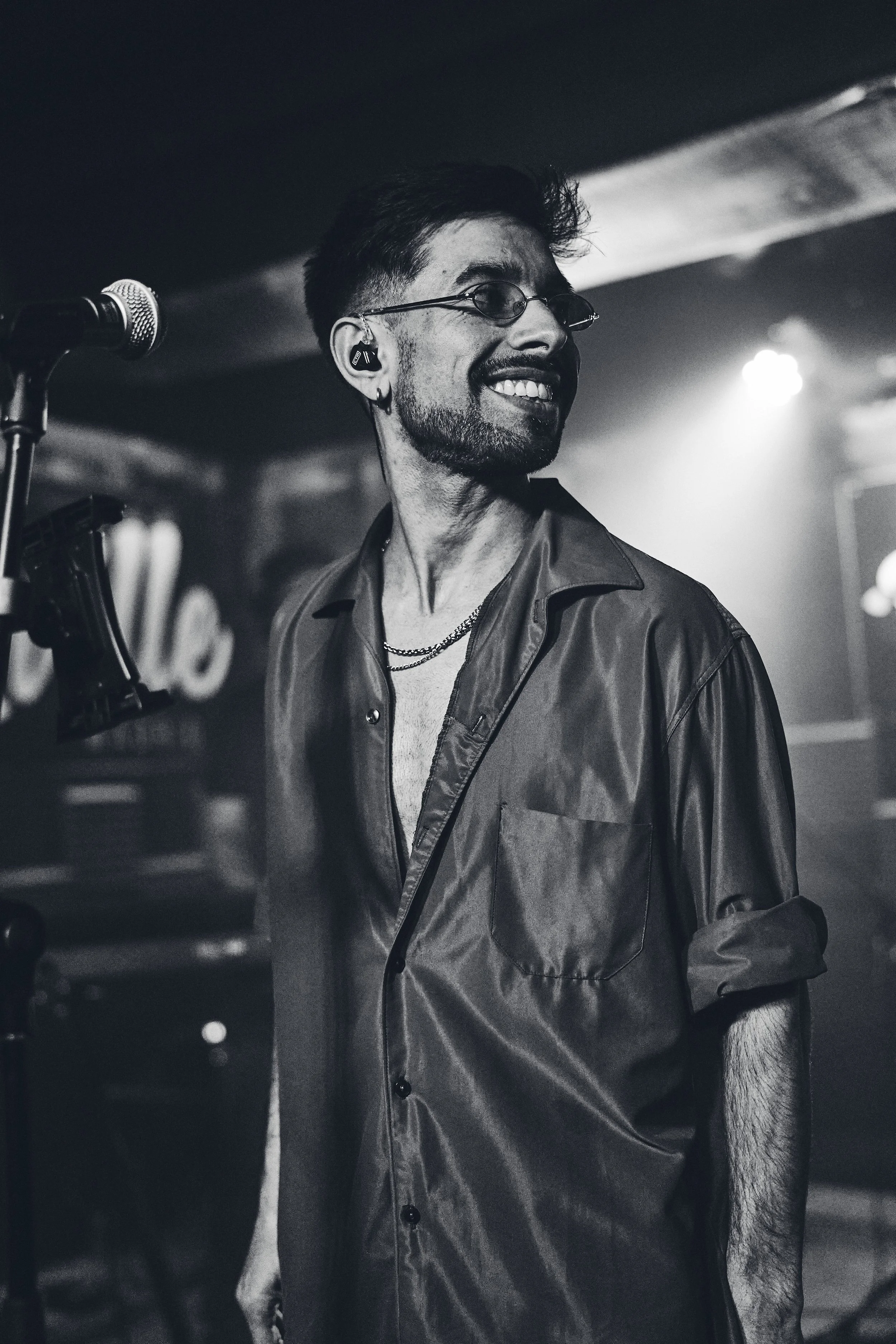 A man with glasses, facial hair, and earrings is smiling on stage during a performance, wearing a shiny button-up shirt with the sleeves rolled up.