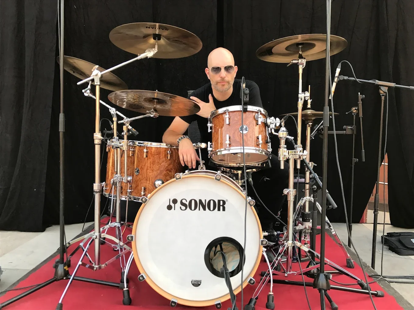 A man with sunglasses sitting at a drum set on a red carpet against a black backdrop.
