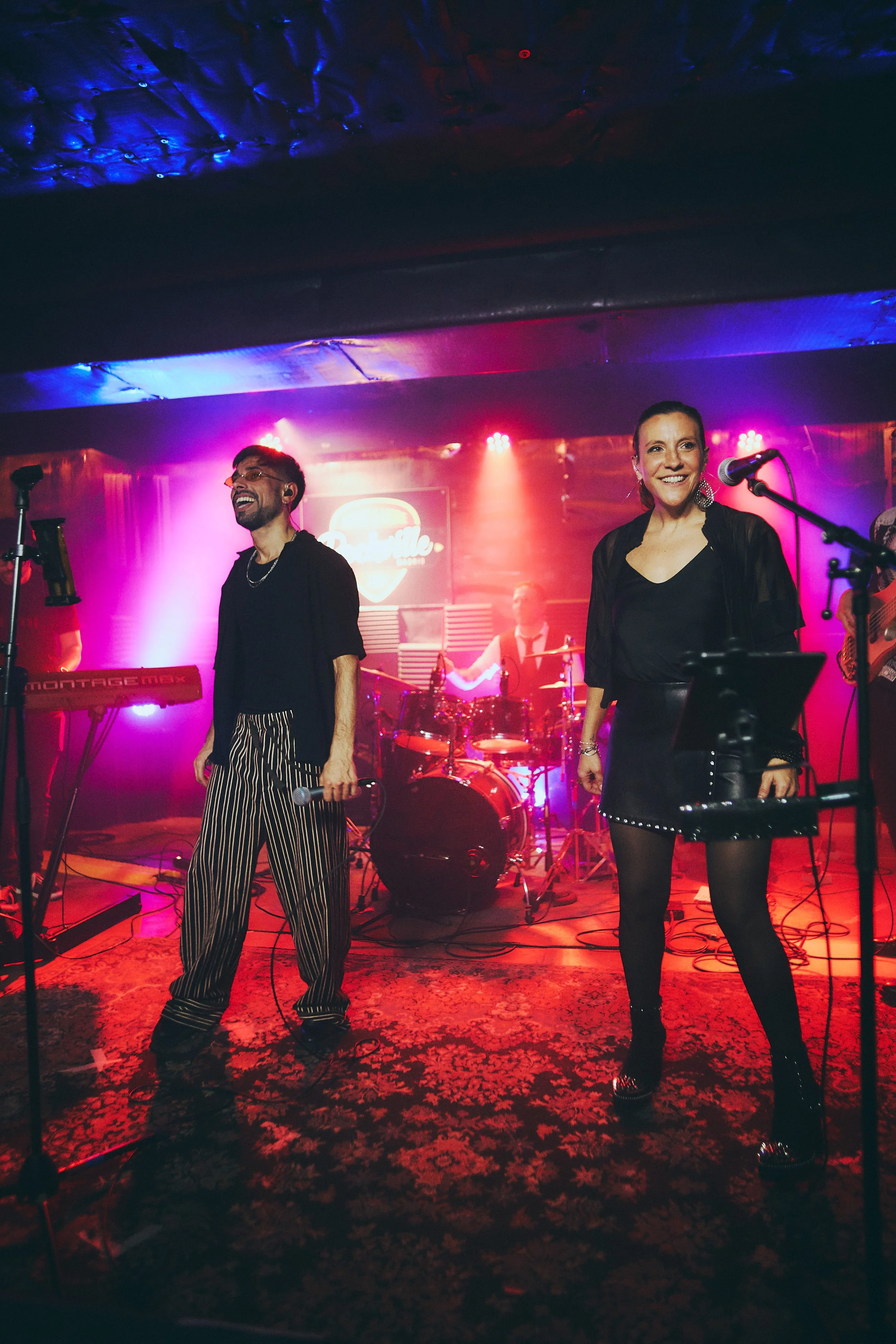 Live band performing on stage with colorful lighting, featuring two singers at the front, one man and one woman, with a drummer and guitarist in the background.