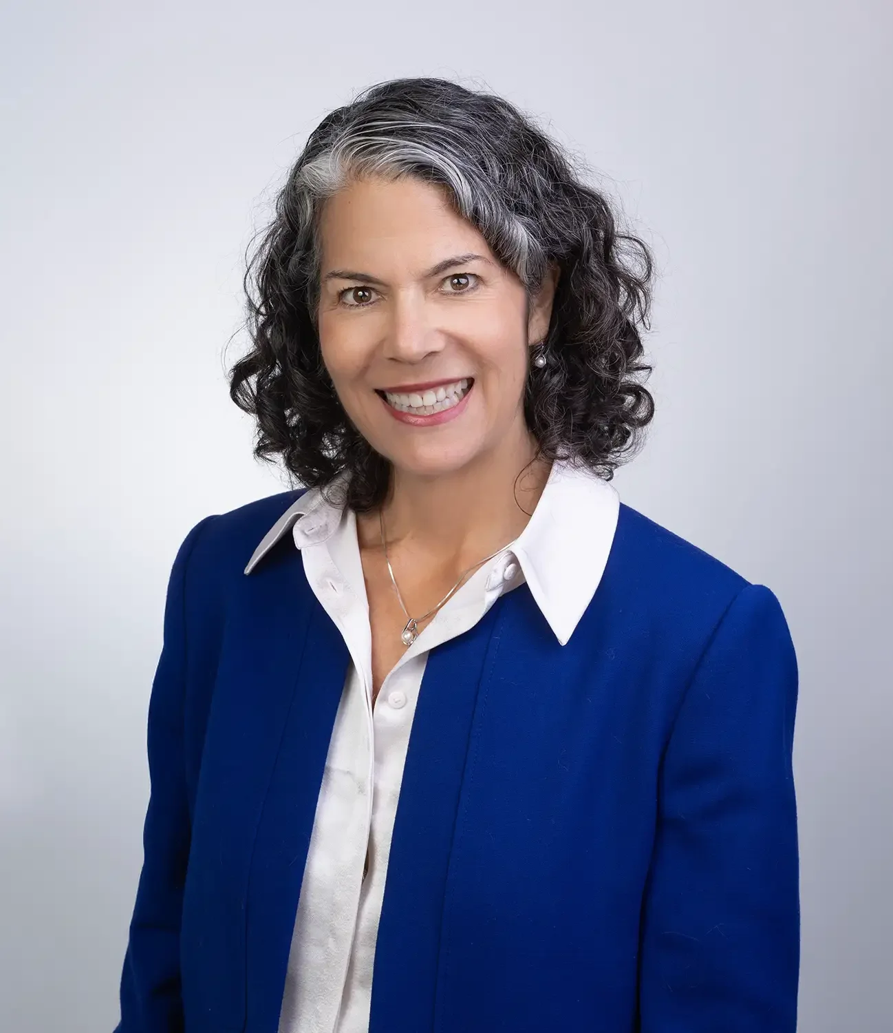 A woman with curly salt-and-pepper hair smiling, wearing a white collared shirt and a blue blazer, against a plain light gray background.