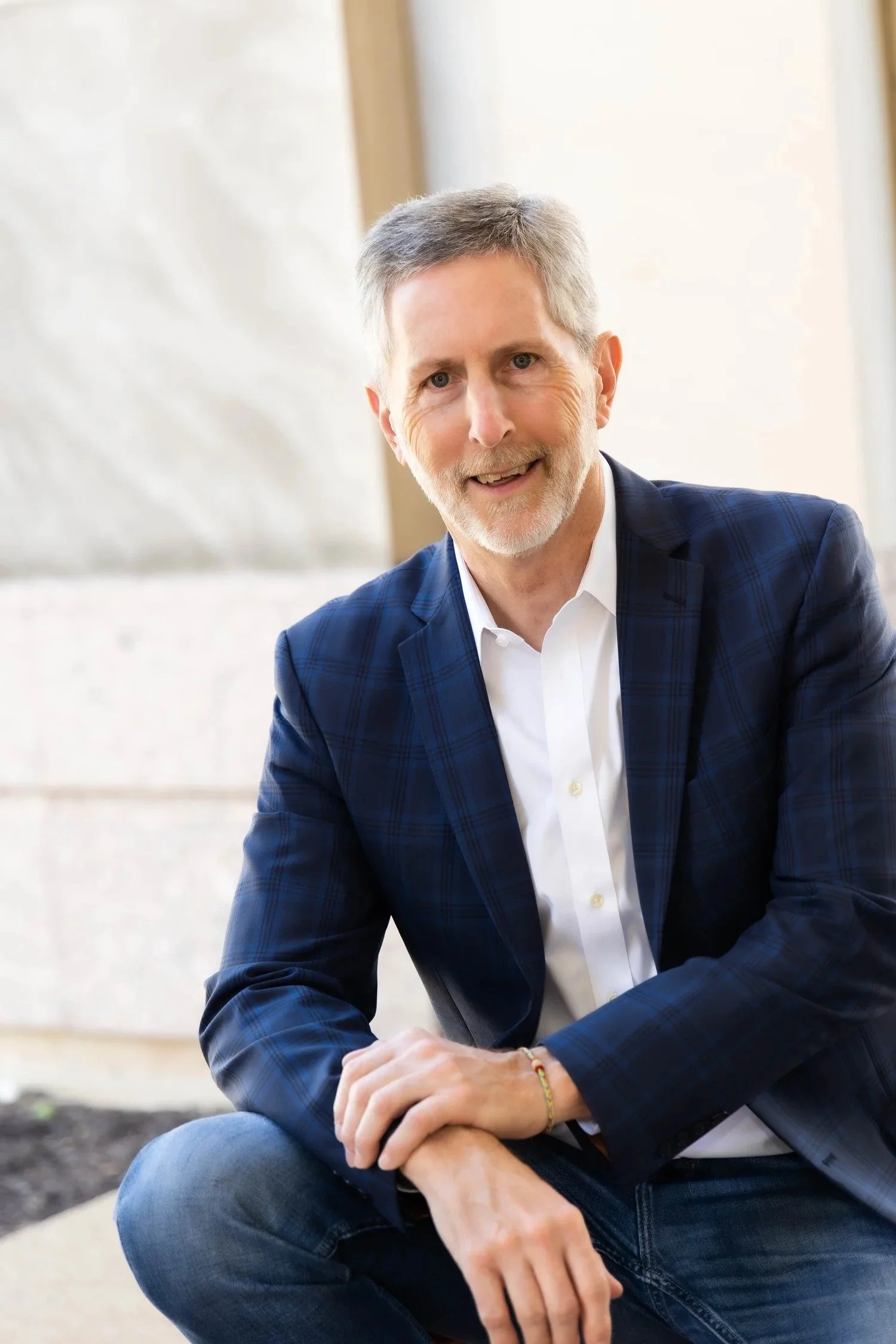 A middle-aged man with gray hair and a beard, dressed in a dark blue plaid blazer and white shirt, sitting outdoors near a stone building with large windows, smiling at the camera.