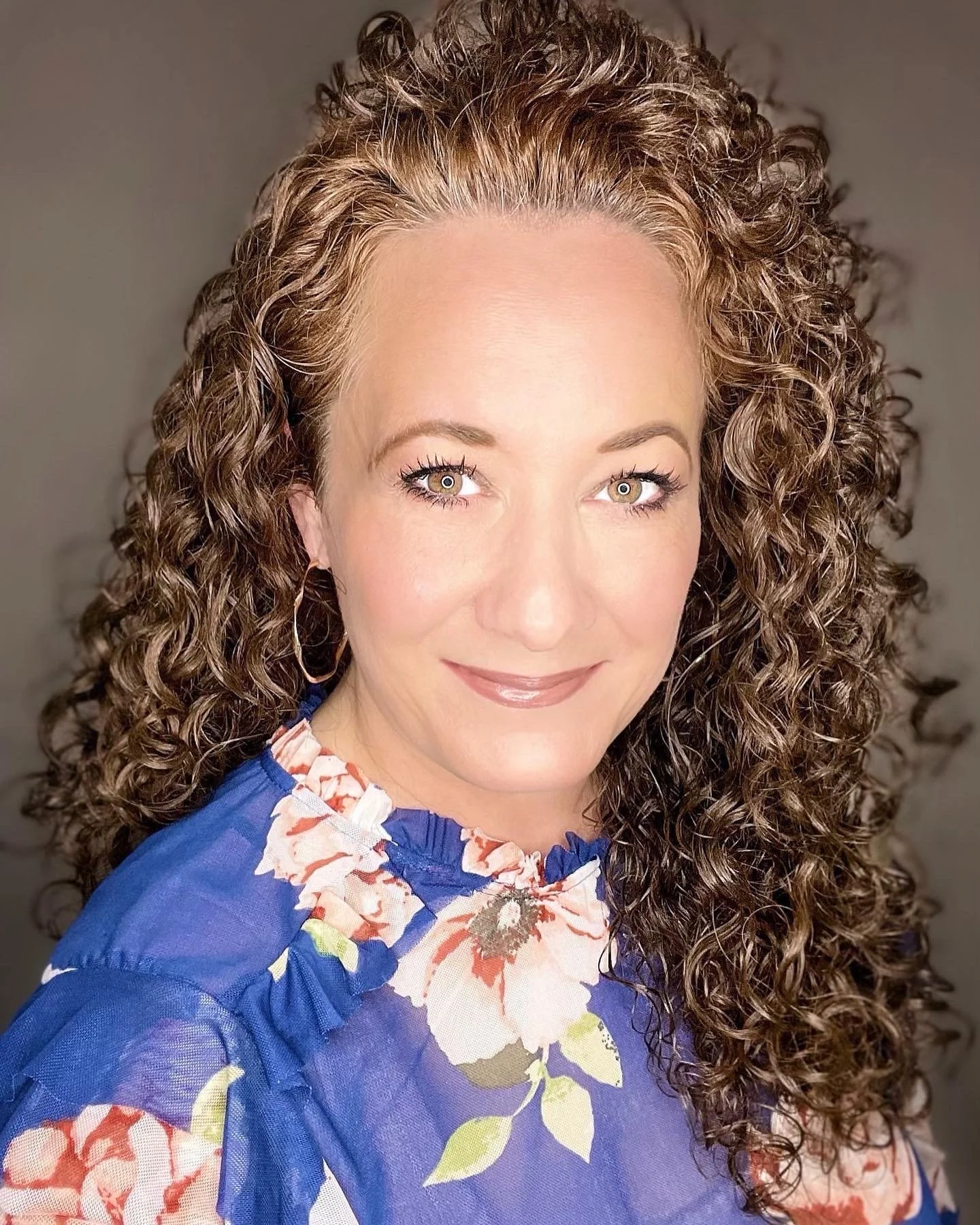 Close-up portrait of a woman with curly brown hair, wearing a blue floral top, smiling.