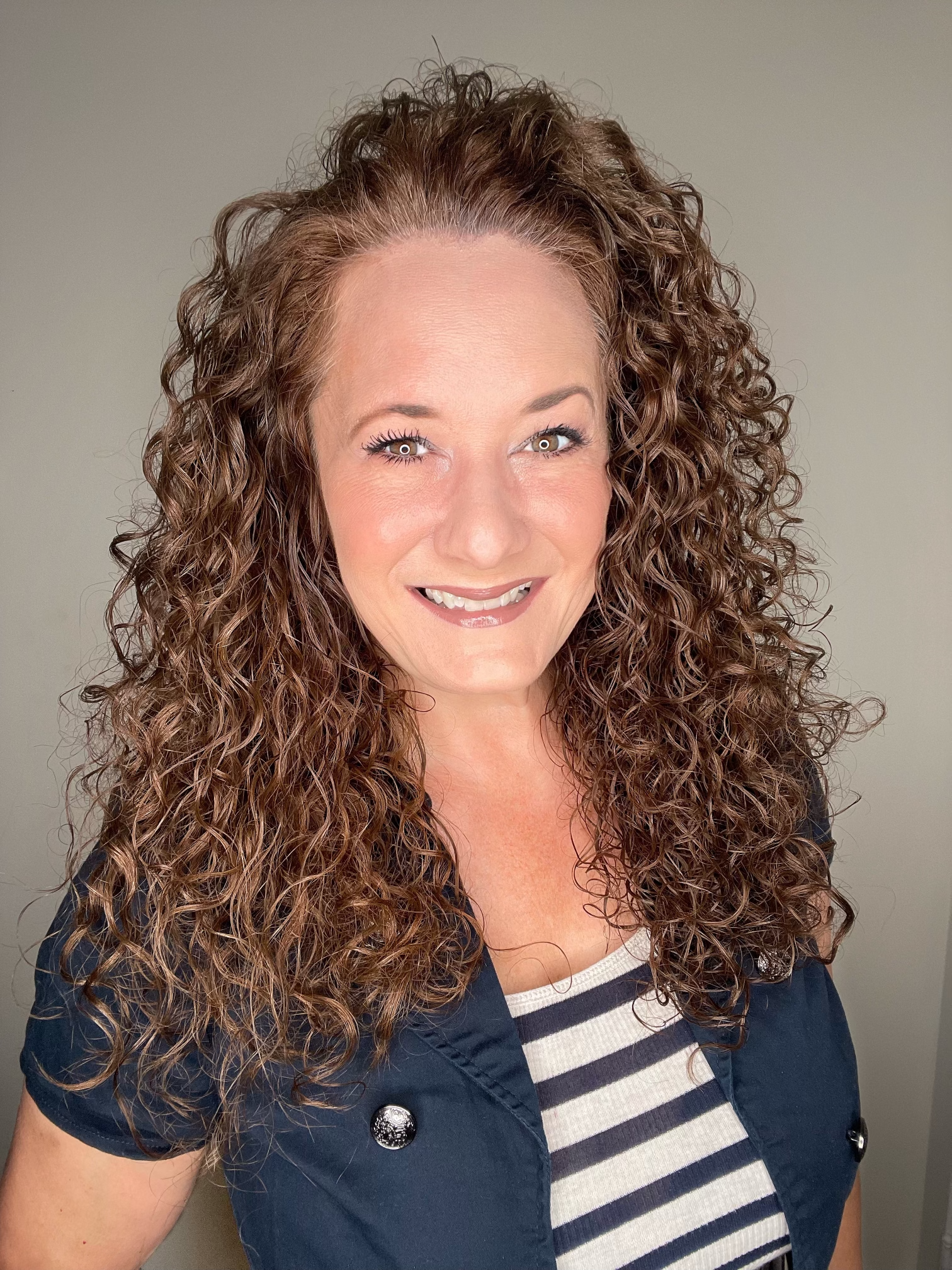 A woman with curly reddish-brown hair smiling at the camera, wearing a black jacket and a striped shirt, against a plain background.
