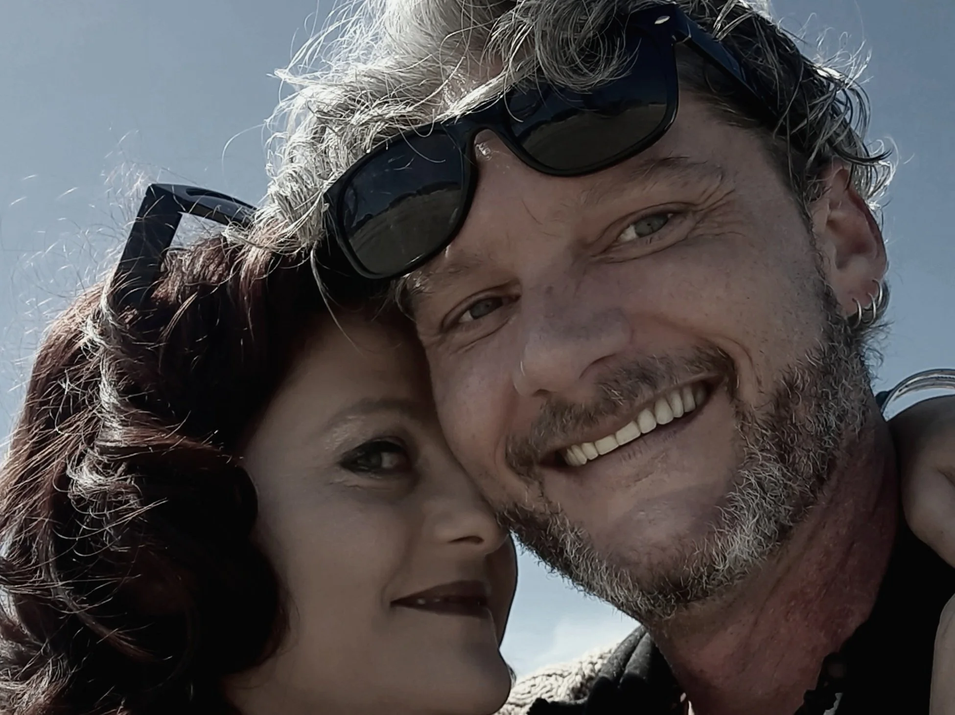 Close-up of a smiling man with sunglasses on his head and a woman with dark hair and makeup, both close together, outdoors with a cloudy sky background.