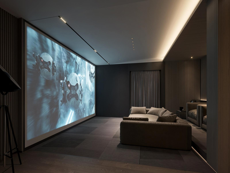 A modern home theater room with a large wall-mounted screen showing a scene from a sci-fi or animated film with robots. The room features a beige sectional sofa with cushions, dark walls, and minimal lighting.