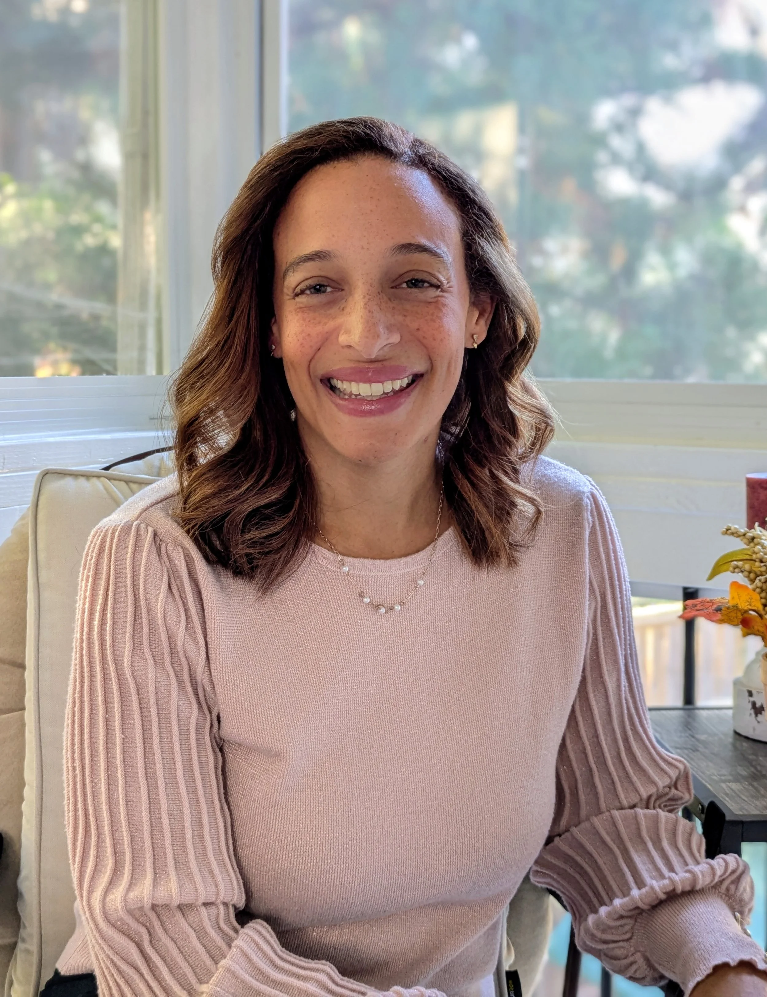 A woman with curly brown hair and freckles smiling indoors near a window with natural light, wearing a light pink sweater and jewelry.