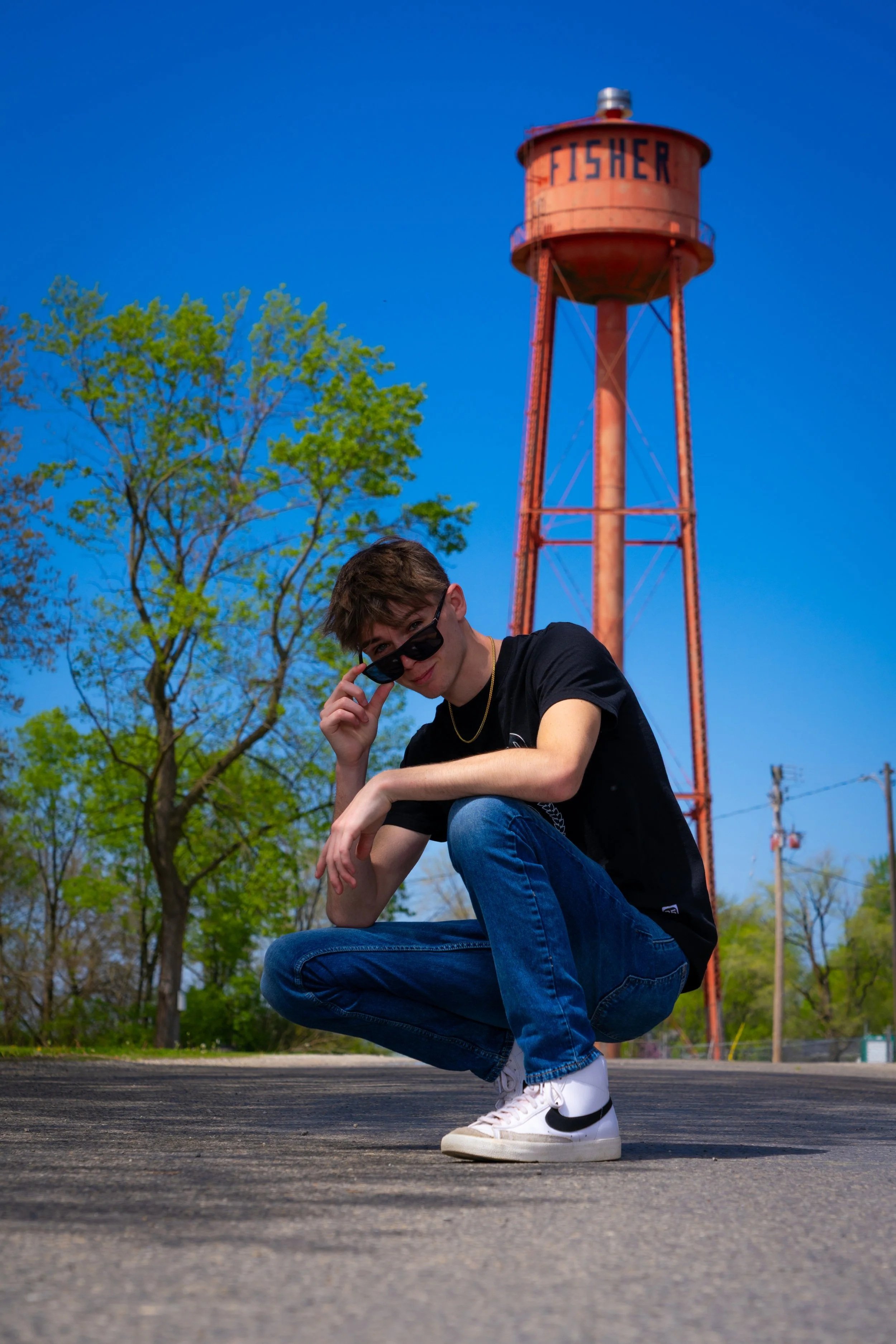 A young man squatting on a paved surface, wearing a black t-shirt, blue jeans, white Nike sneakers, and black sunglasses, looking at the camera with a playful expression. In the background, there is a tall red water tower with the word 'FISHER' painted on it, green trees, and utility poles under a bright blue sky.