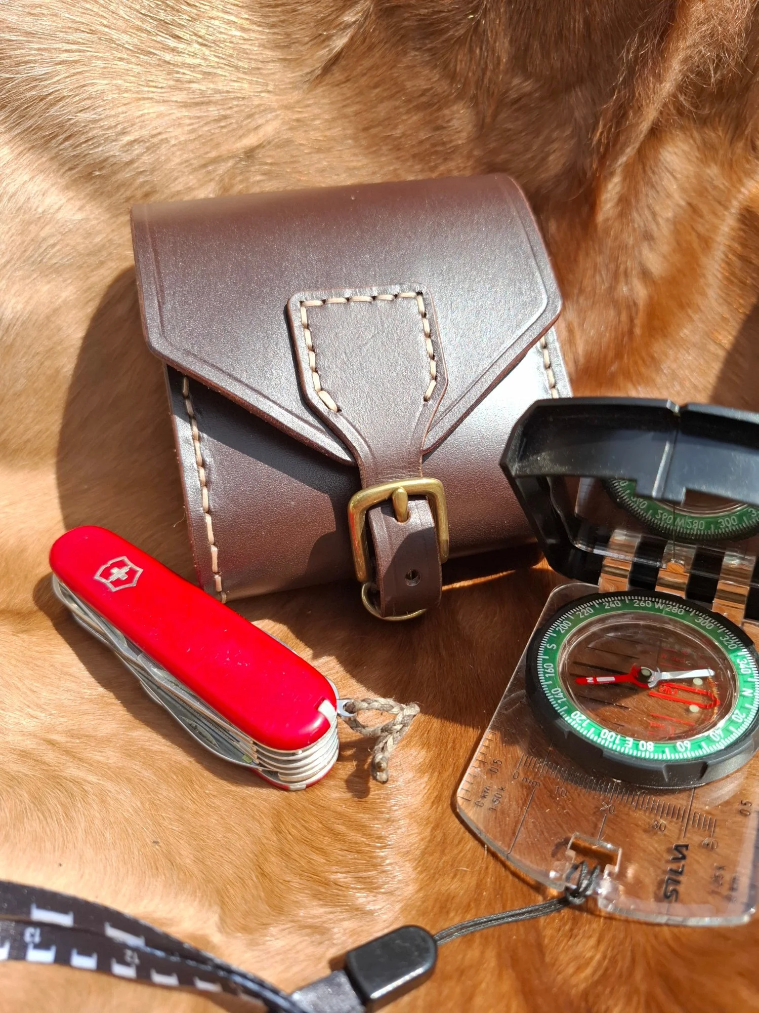 Close-up of a brown leather bag, a red Swiss Army knife, a black and green compass, and a transparent protractor on a wooden surface with fur texture.