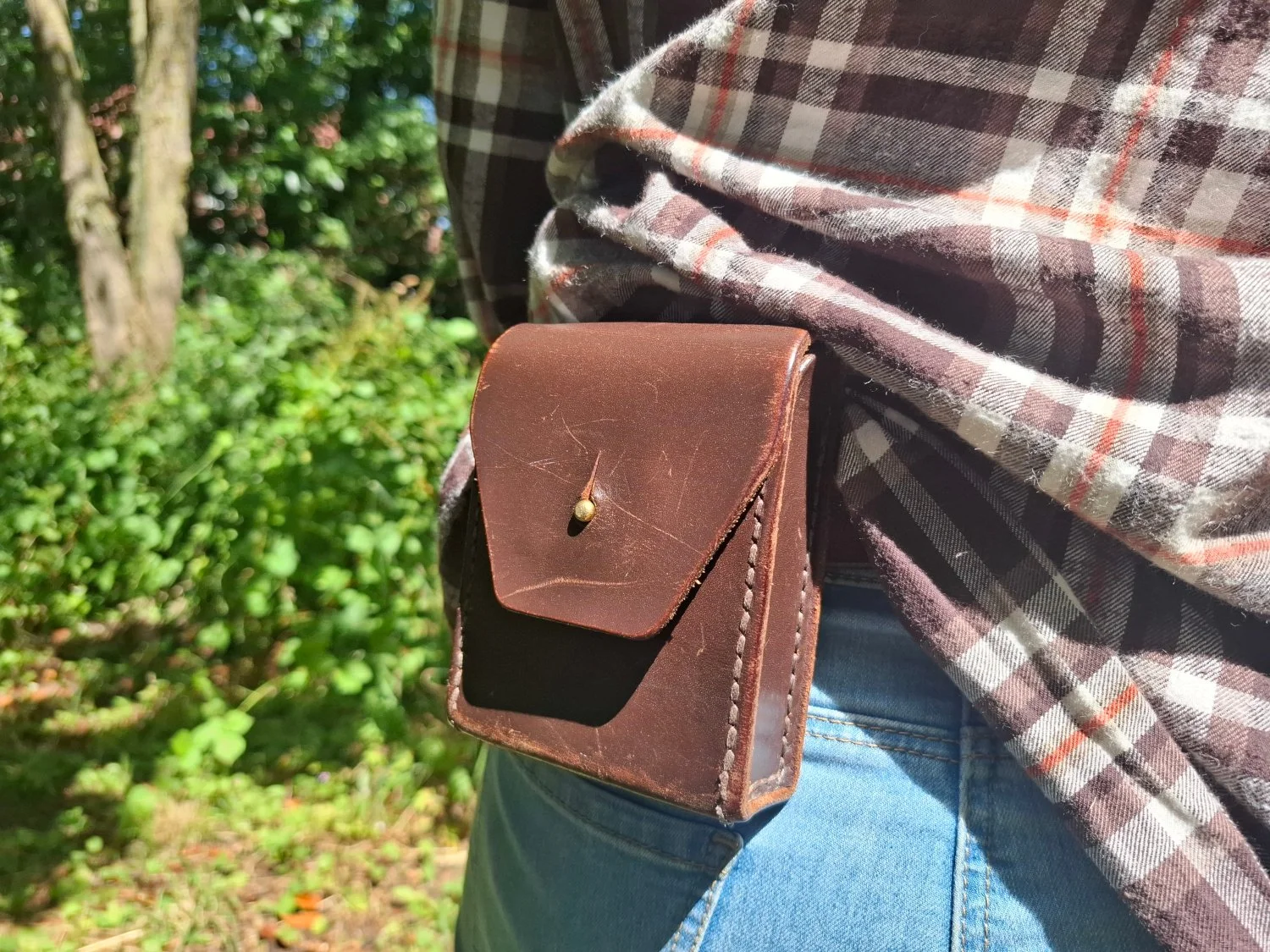 Close-up of a person's hip and waist, with a leather holster or pouch attached to their belt, partially covering blue jeans and a plaid shirt. The background shows green trees and foliage, indicating an outdoor setting.