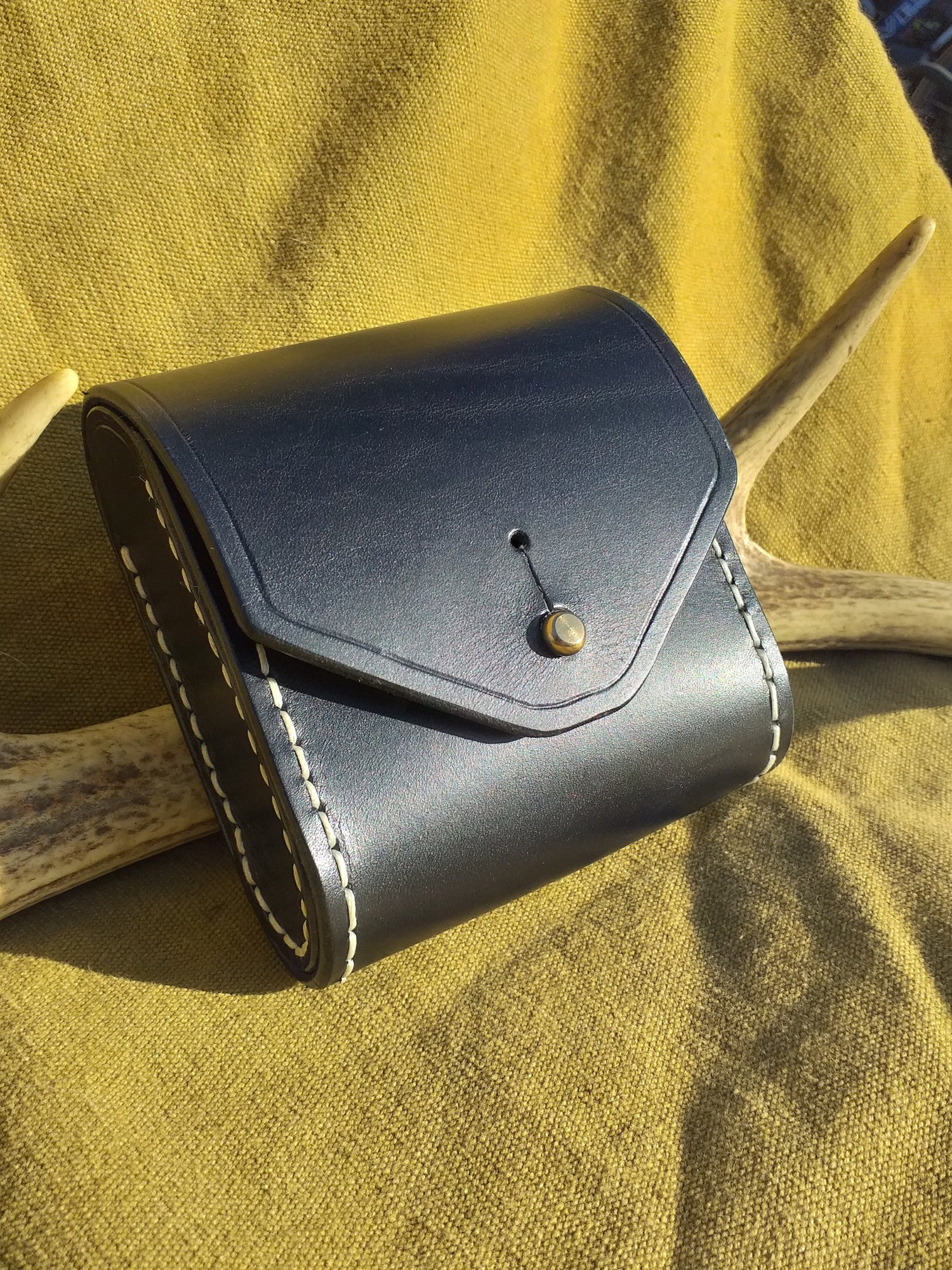 A black leather purse with white stitching, placed on a yellow textured fabric surface, with a wooden object and part of a chair visible in the background.