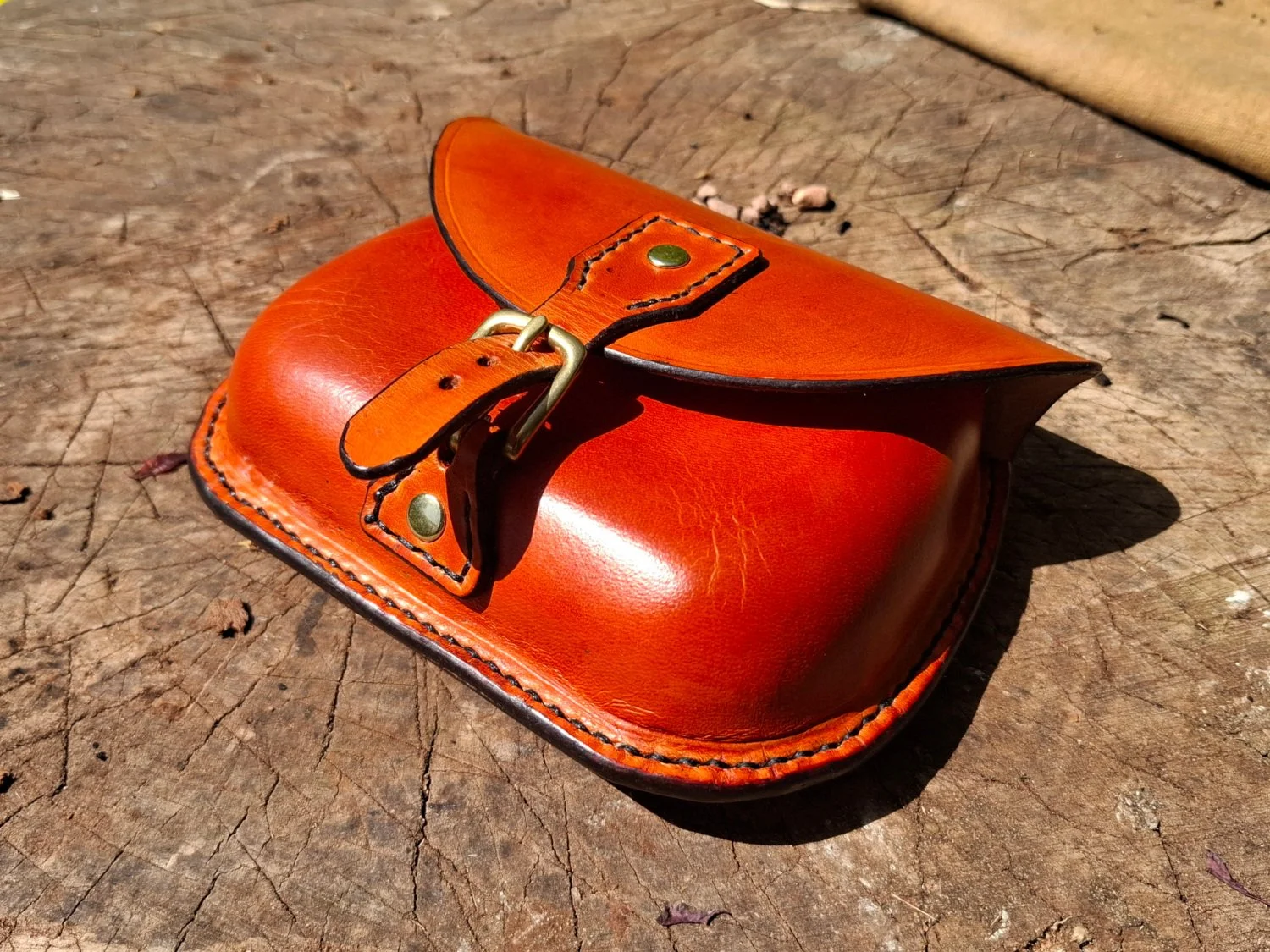 A small, orange leather pouch with a flap and buckle closure resting on a weathered wooden surface.