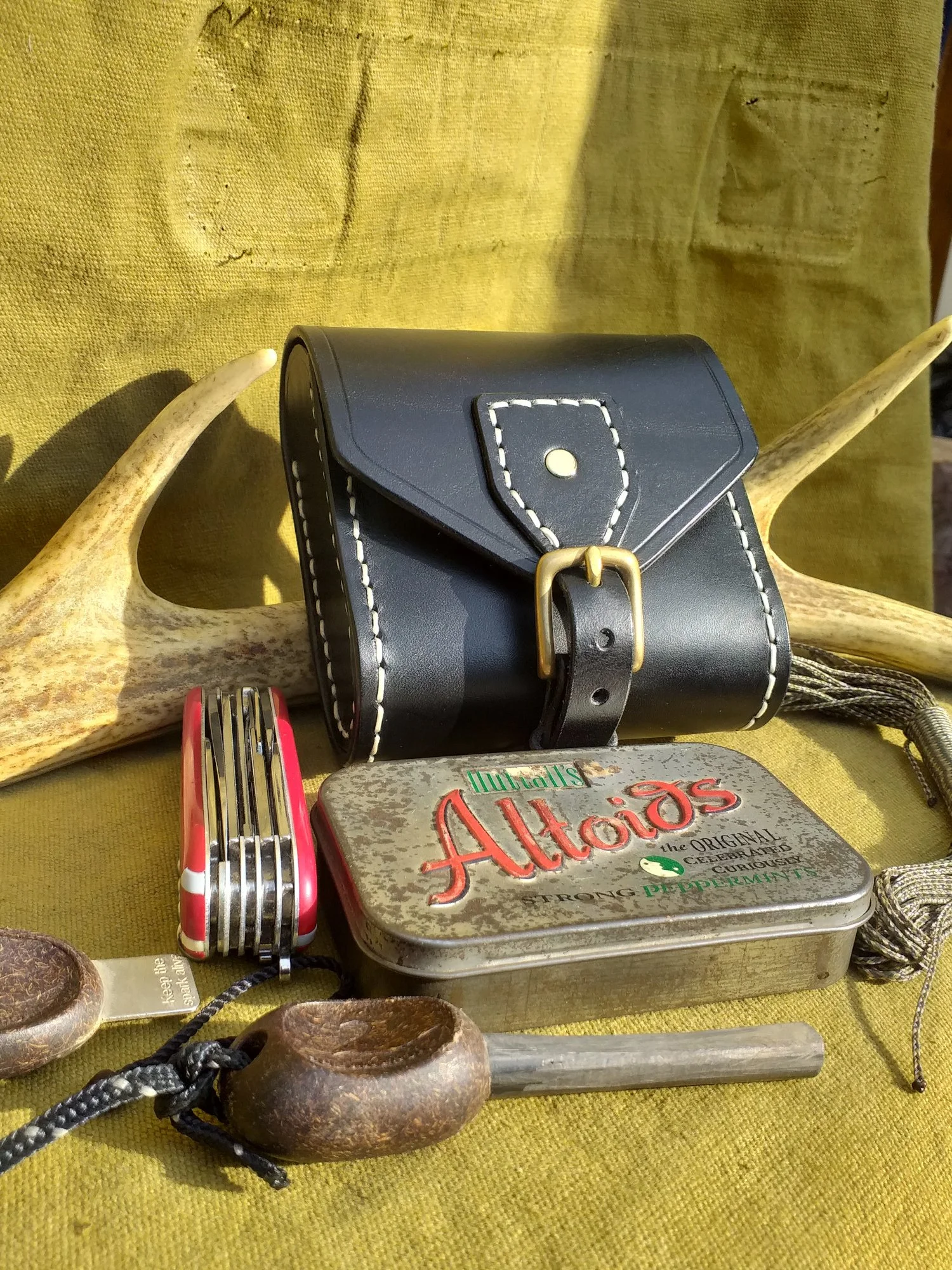 A collection of vintage items including a black leather camera bag with white stitching and brass buckle, a red Swiss Army knife, a metal Altoids gum tin, a wooden ladle, wind chimes, an antler, and a yellow fabric background.
