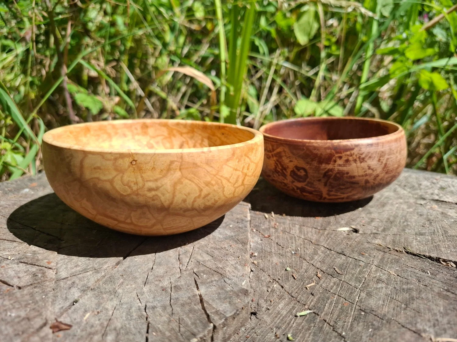 Two wooden field maple bowls, one light-colored and one dark-colored, placed on a wooden stump outdoors with green foliage in the background.