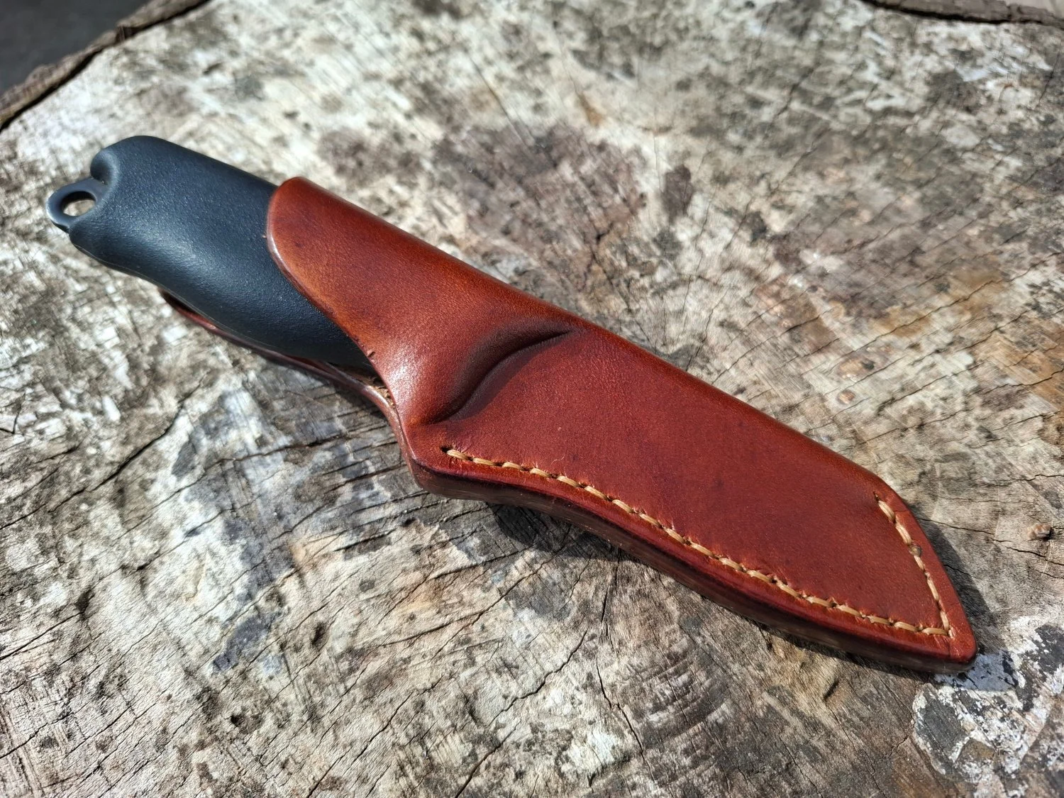 A leather-handled outdoor knife in a brown leather sheath resting on a weathered wooden surface.
