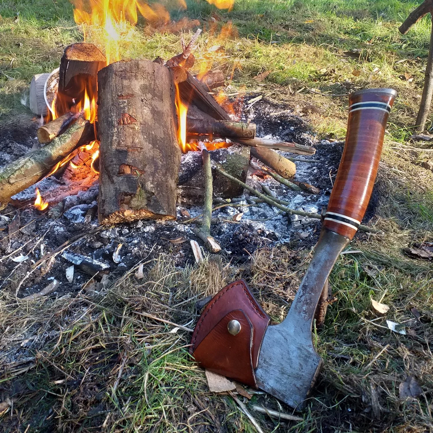 A campfire with burning logs and flames on green grass, an axe with a wooden handle and leather sheath on the ground nearby, and a cup on the right side.