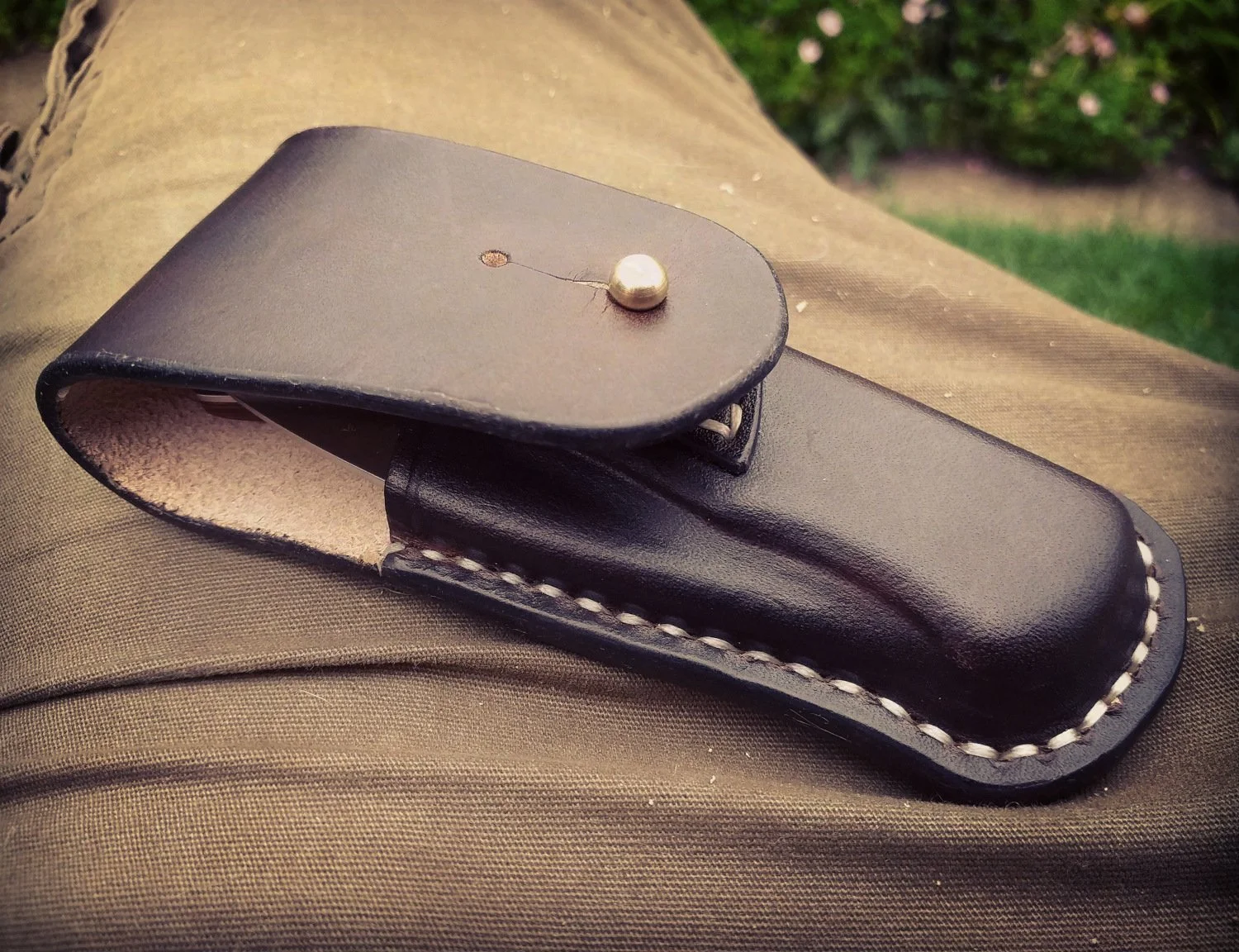 A black leather sheath with a brass button on top, resting on khaki-colored fabric.