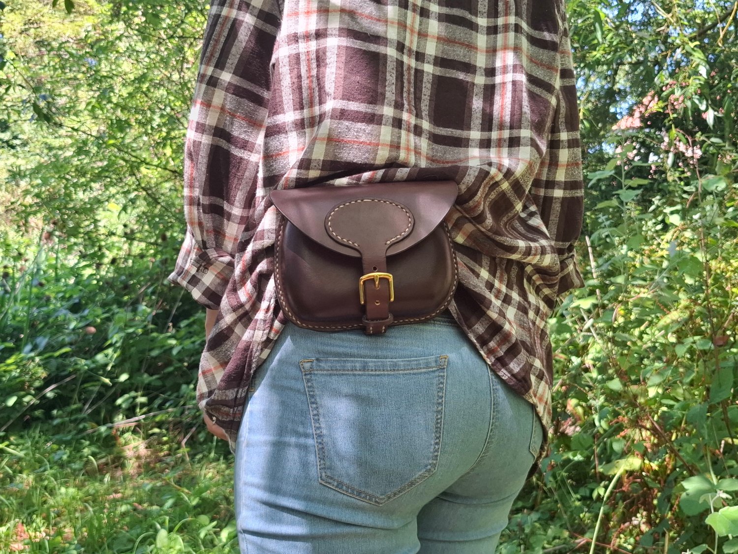 Person wearing a plaid shirt and blue jeans, with a small brown leather belt bag around their waist, standing outdoors in a wooded area.
