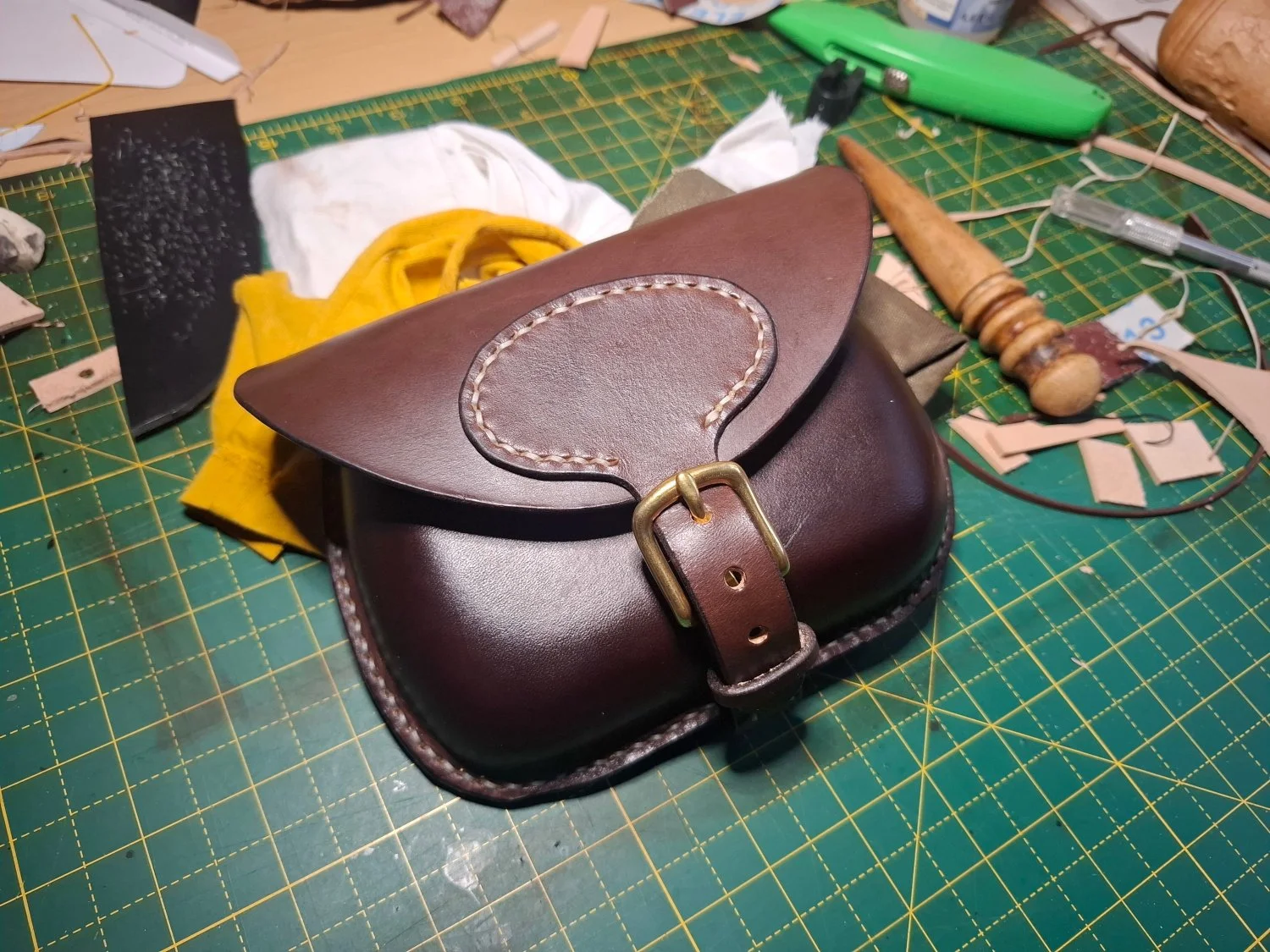A leather handbag on a green cutting mat surrounded by tools and craft materials.