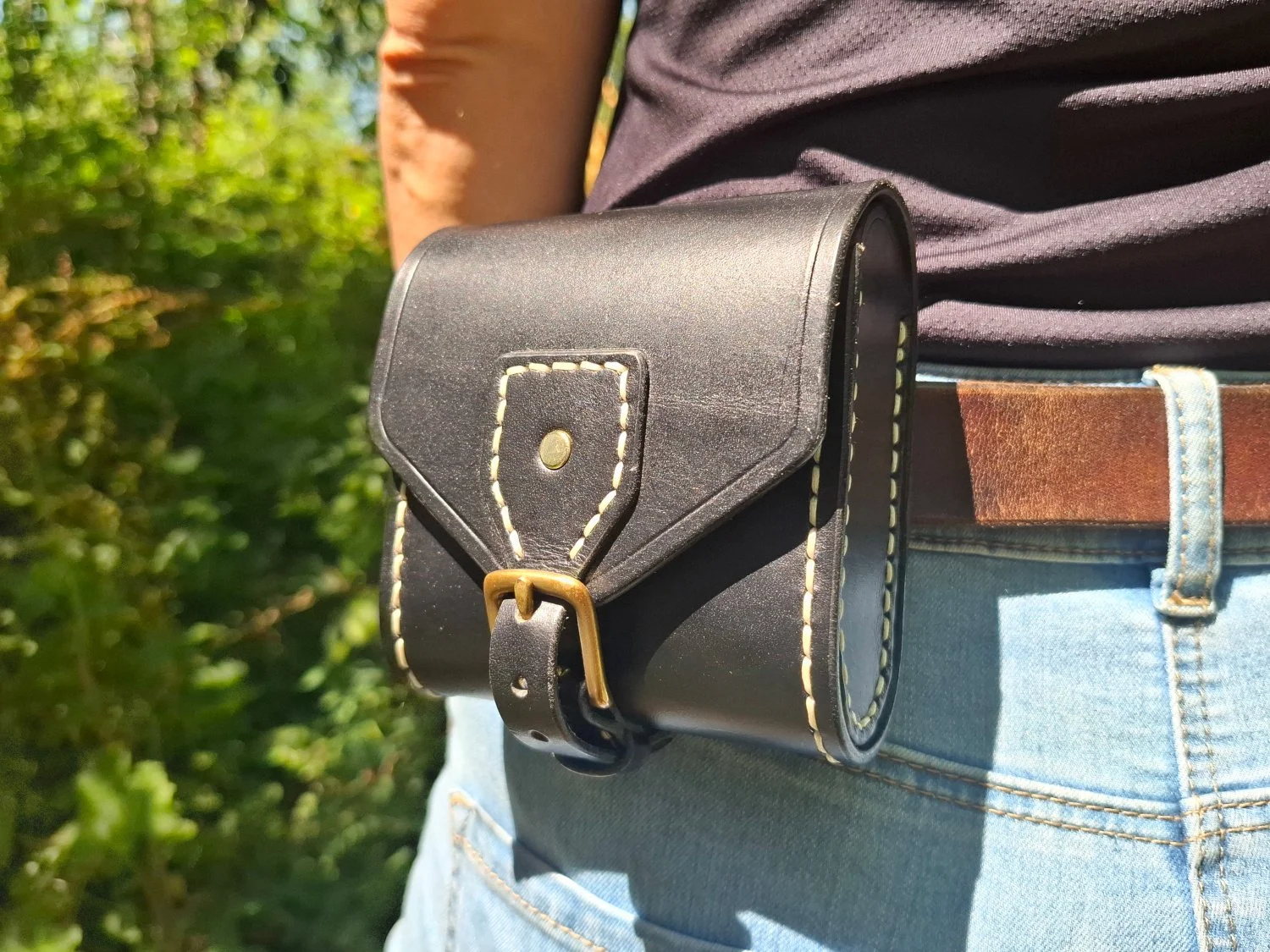 Person wearing light blue jeans, a brown belt, and a dark t-shirt, standing outdoors with green foliage in the background, carrying a small black leather bag with white stitching and a brass buckle.