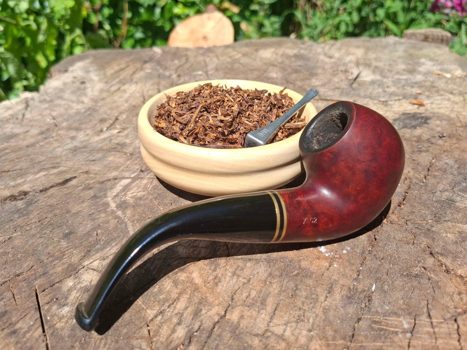 A wooden surface with a bowl of loose tobacco, a metal pipe, and a small scoop, with greenery in the background.