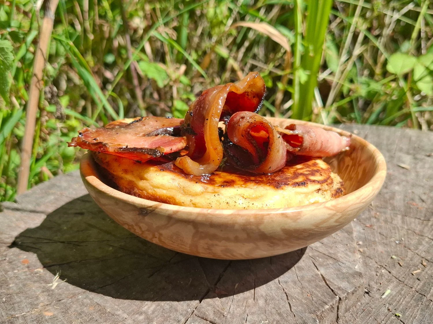 Cooked bacon and onions on a toasted English muffin in a wooden bowl outdoors.