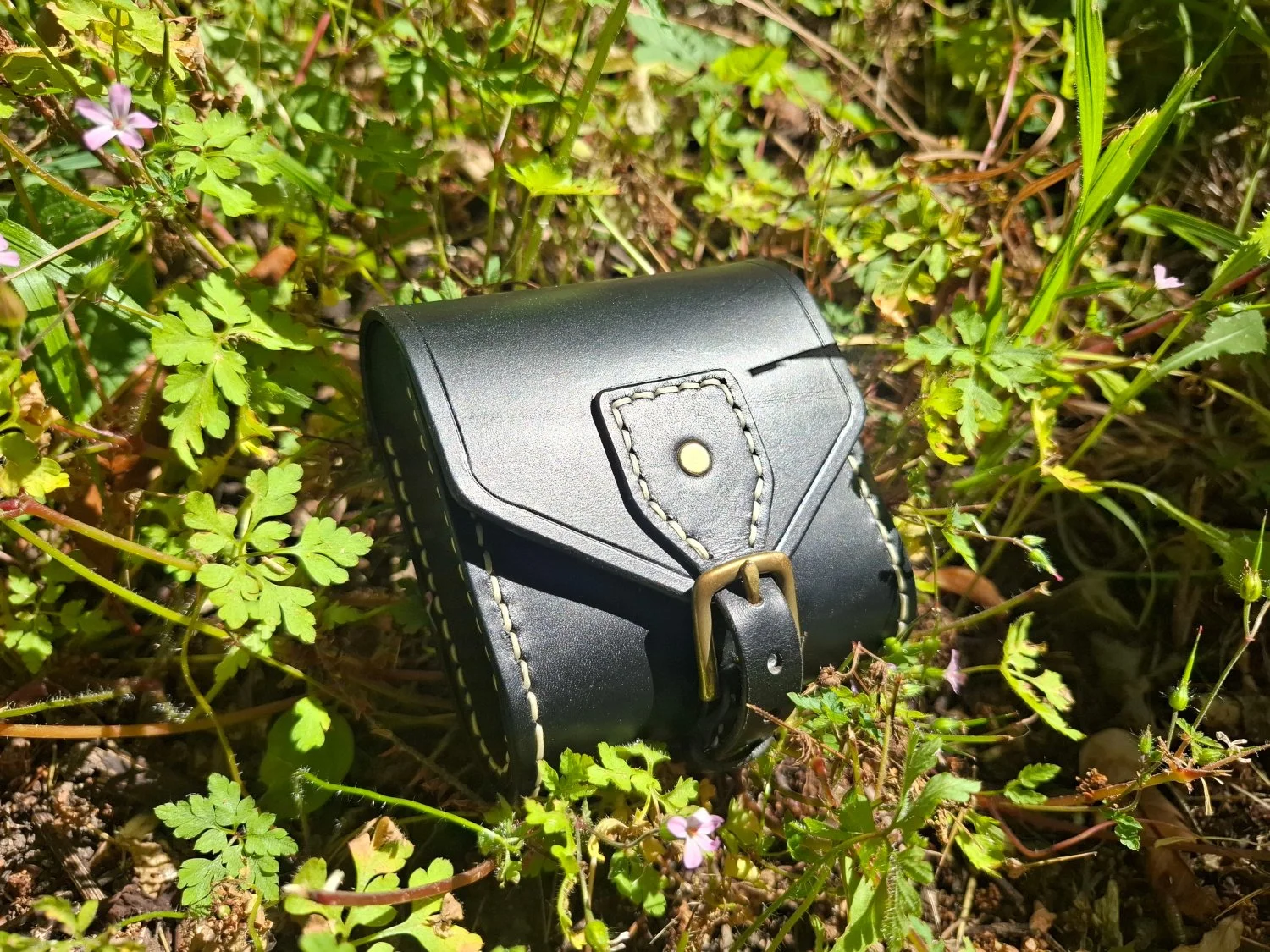 A black leather camera case with beige stitching and a metal buckle is lying on green plants and small pink flowers growing in the ground.