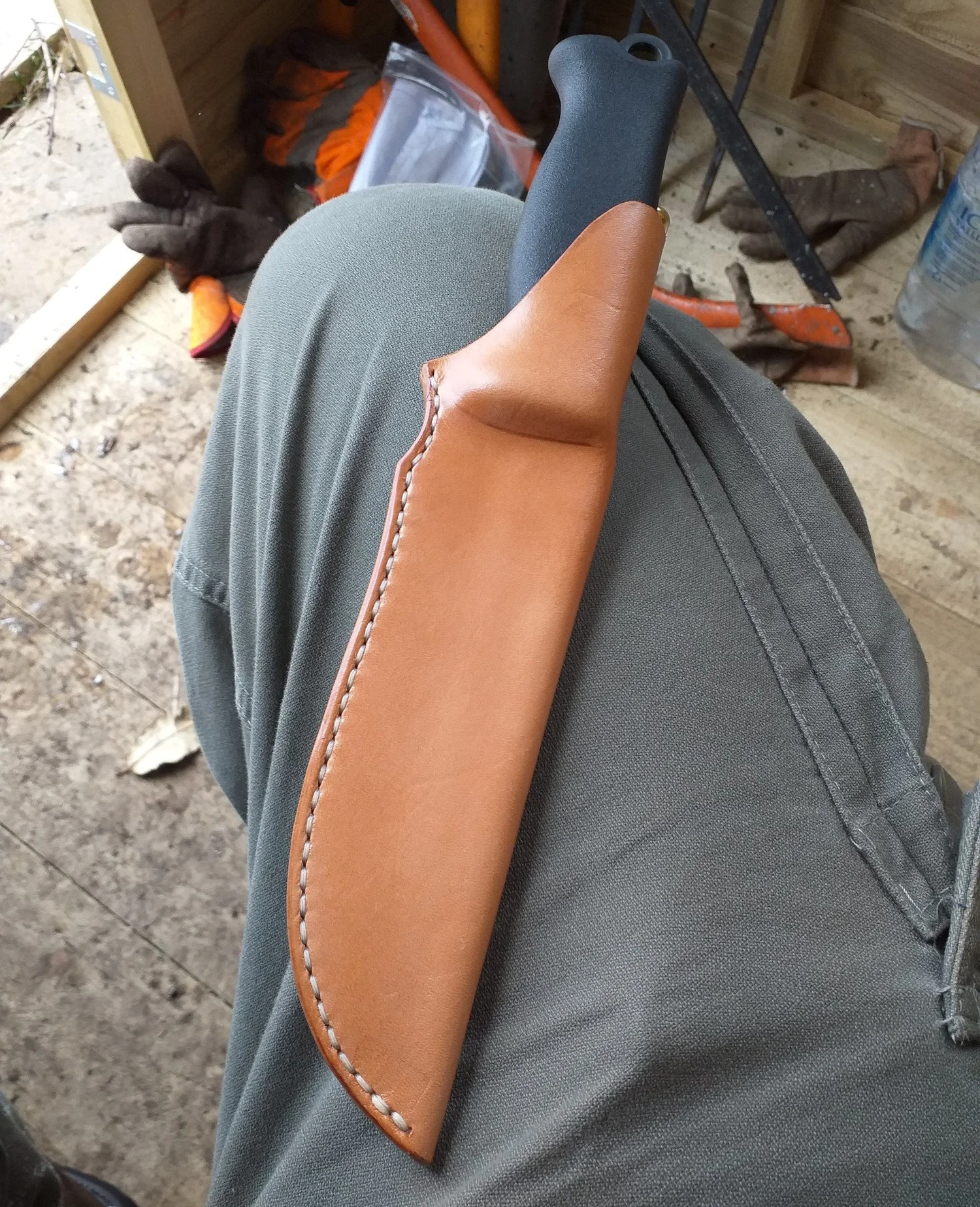 A person sitting outdoors wearing gray pants and holding a brown sheath with a knife handle visible. The background shows dirt, gloves, and gardening tools.
