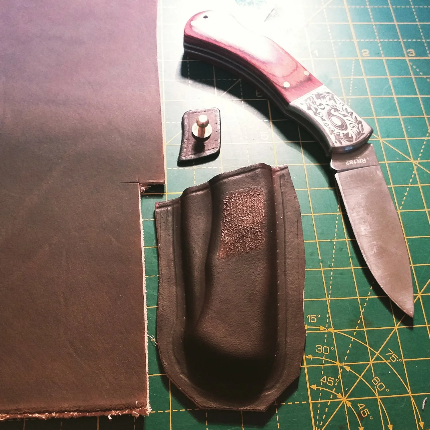 A leather knife sheath, a folding knife with a wooden handle, a small metal pin, and a piece of leather on a green cutting mat with grid lines.