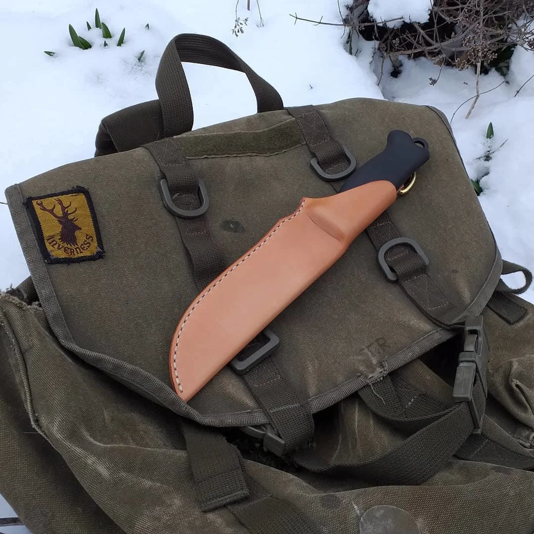 A beige-handled knife with a black blade sheath resting on a green backpack outside on snow.