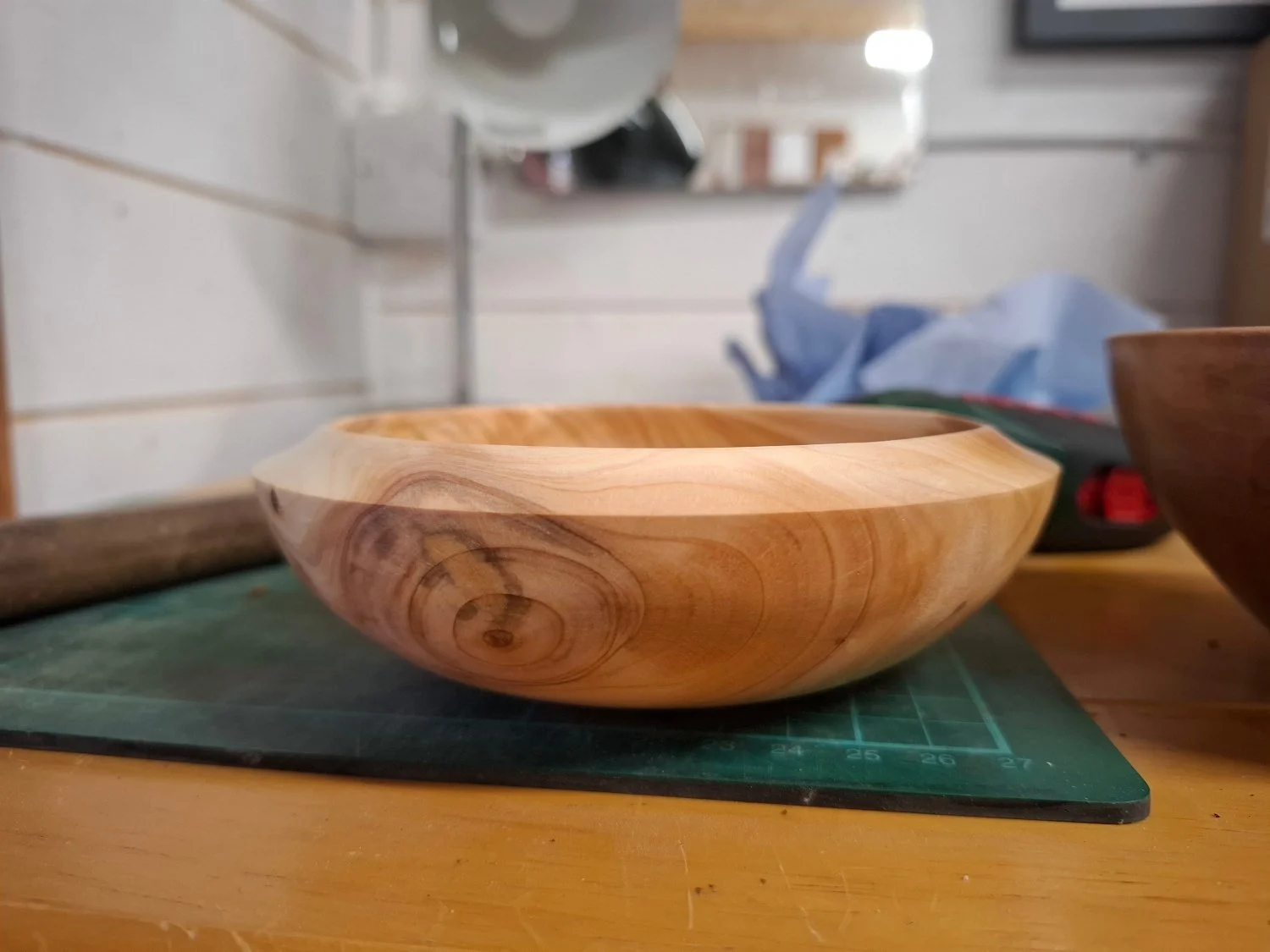 A wooden bowl with a smooth finish and natural grain pattern sits on a green cutting mat on a wooden table. In the background, there are blurred objects including a mirror, containers, and a blue paper bag.