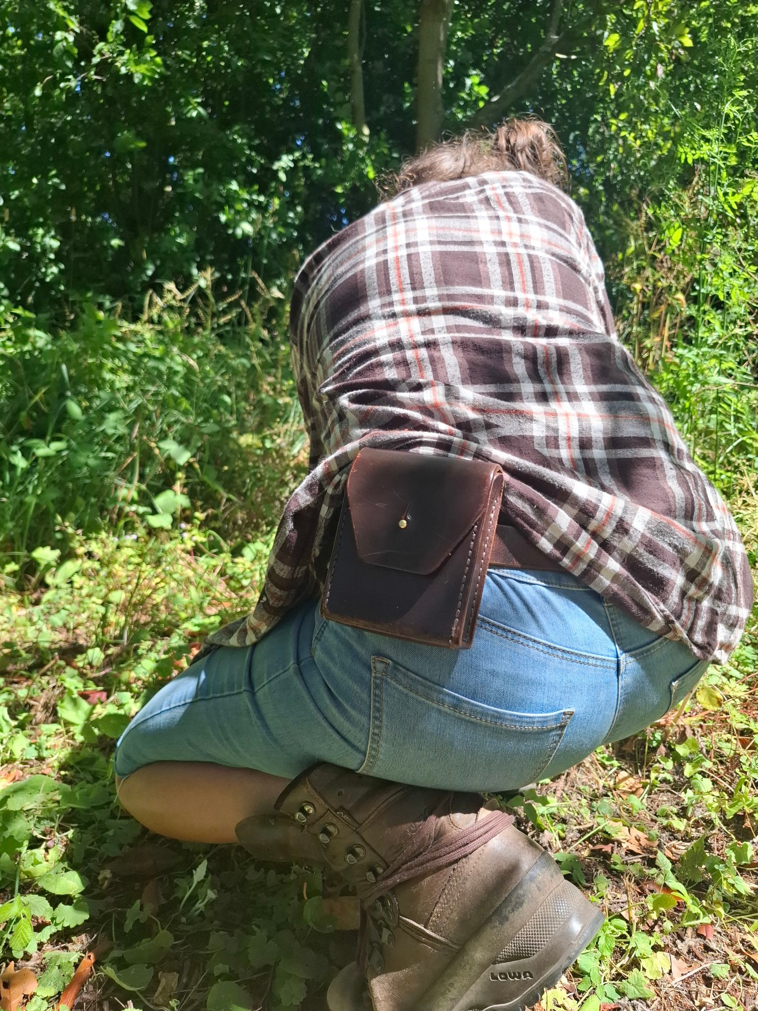 A person wearing a plaid shirt, blue jeans, and hiking boots, kneeling or crouching among green plants in a forested area.