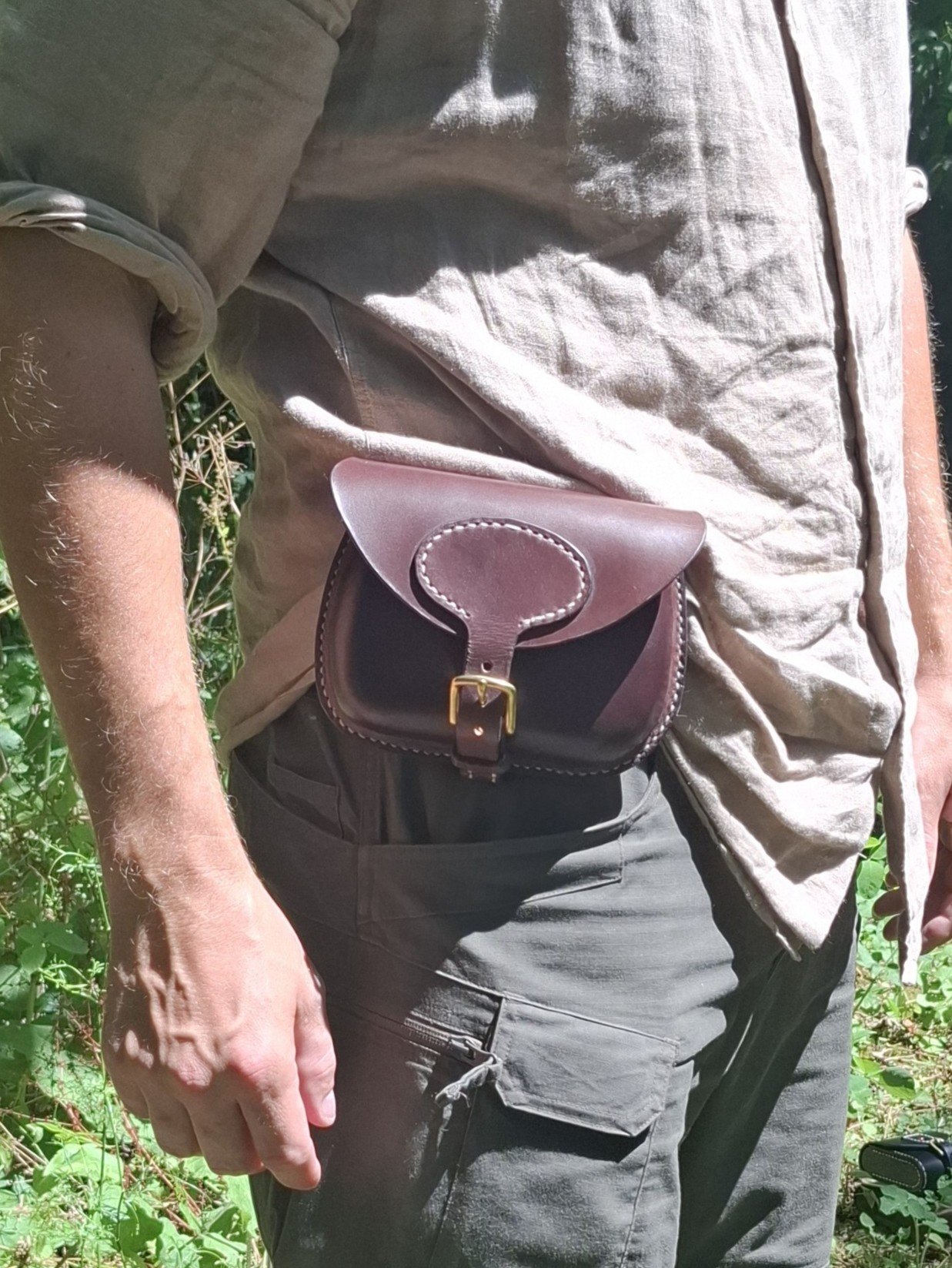 Person wearing a beige shirt and cargo pants standing outdoors with a small brown leather crossbody bag.