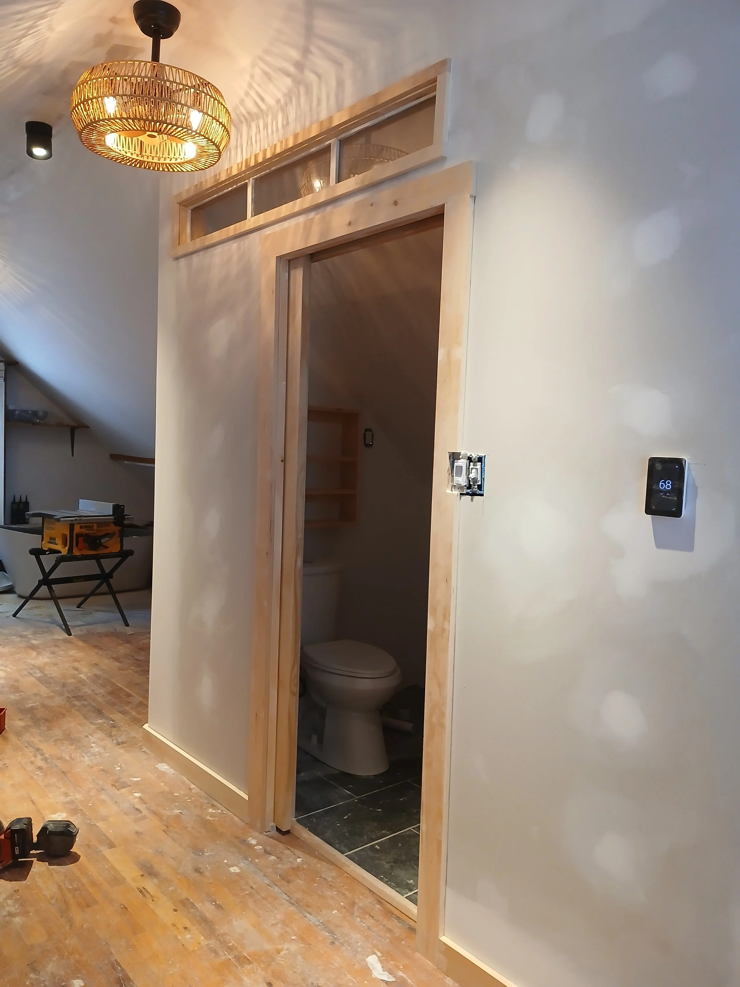Partial view of a room under construction, with a small bathroom with a toilet visible inside. The bathroom has a slate tile floor, and the doorway is framed with unfinished wood. There is a wall-mounted thermostat displaying 68 degrees and a light s